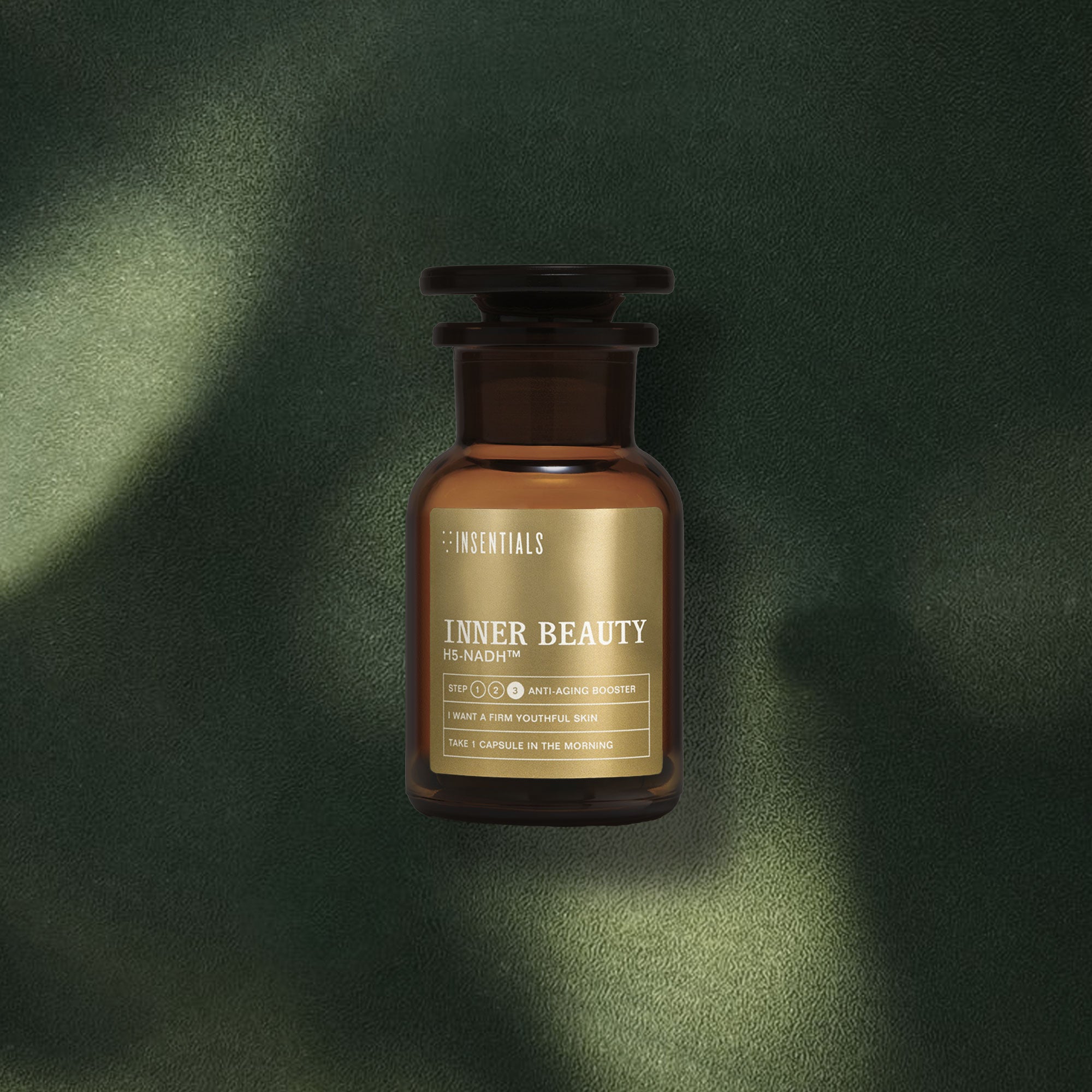 Een amberkleurige glazen fles met het etiket „Inner Beauty H5-NADH™ (huidverjonging op celniveau)” van Insentials staat op een groen oppervlak met structuur, badend in gevlekt zonlicht dat de belofte van het product benadrukt: het ondersteunen van cellulaire energie en het bevorderen van een jeugdige huid op de lange termijn.