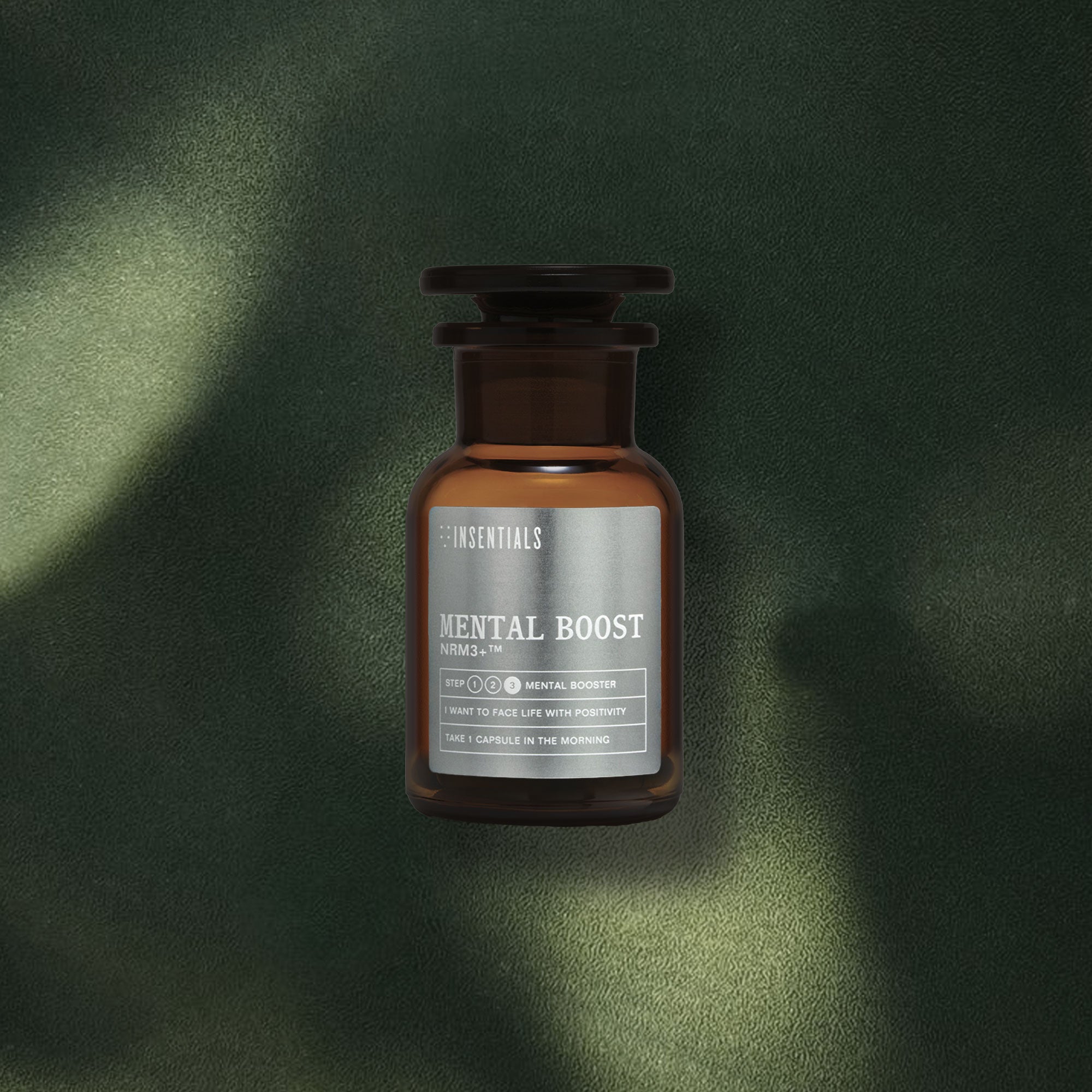 Een bruine glazen fles met het etiket „NRM3+ (Mental Boost) – Focus & Concentratie” van Insentials – met neuro-regeneratieve paddenstoelen zoals Lion’s Mane en andere noötropica – staat op een donkergroen, gestructureerd oppervlak dat door een dramatisch spel van licht en schaduw wordt geaccentueerd.