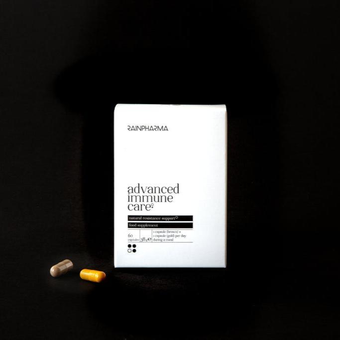 Een witte doos "Advanced Immune Care" van RainPharma staat op een zwarte achtergrond, met gele en beige capsules ernaast. Deze krachtige formule ondersteunt het immuunsysteem en bevordert een gezonde huid.