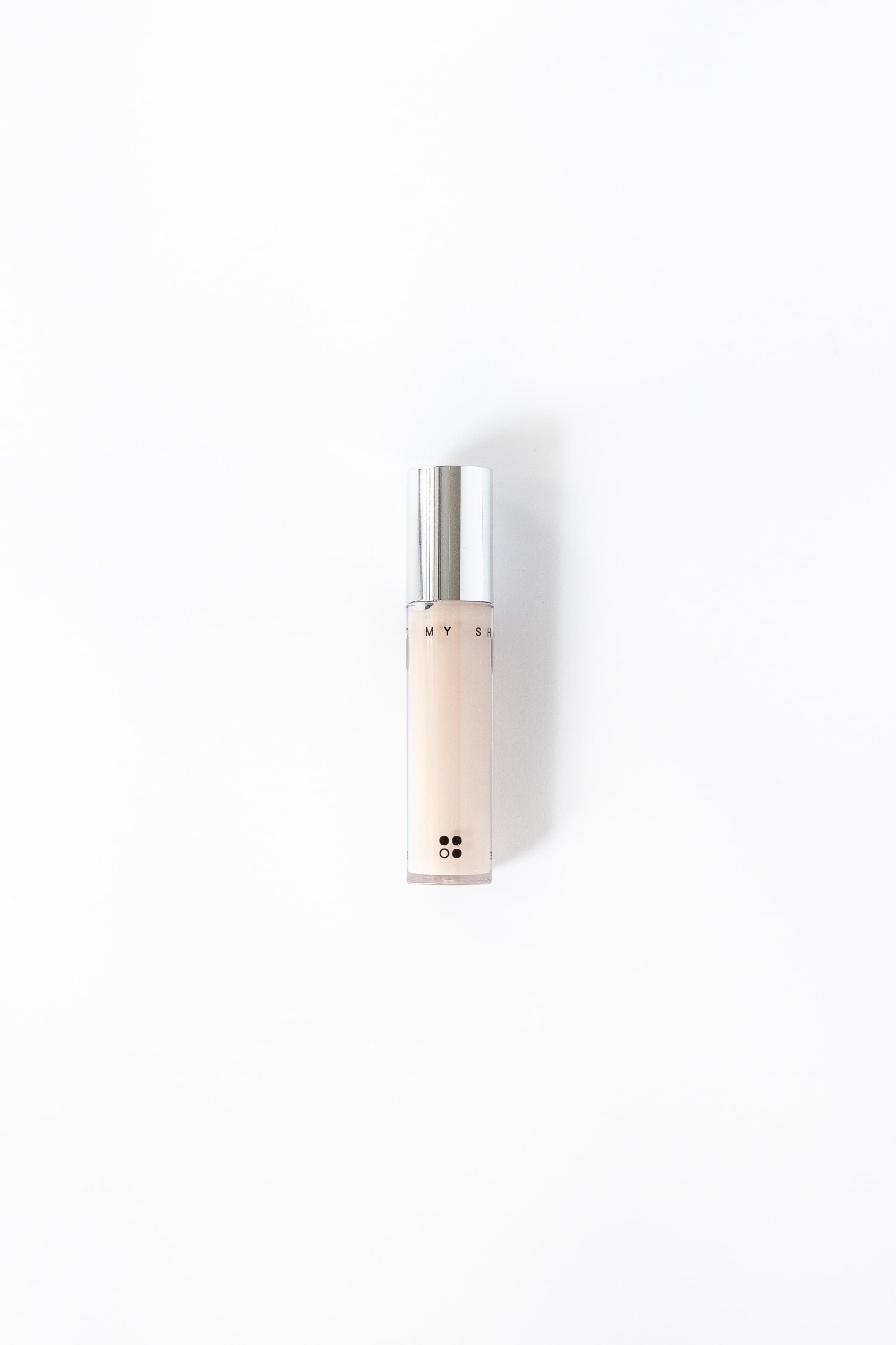 Een klein, cilindervormig flesje beige "Light my Shadow" veganistische concealer van RainPharma, met een glanzende zilveren dop, steekt elegant af tegen een witte achtergrond.