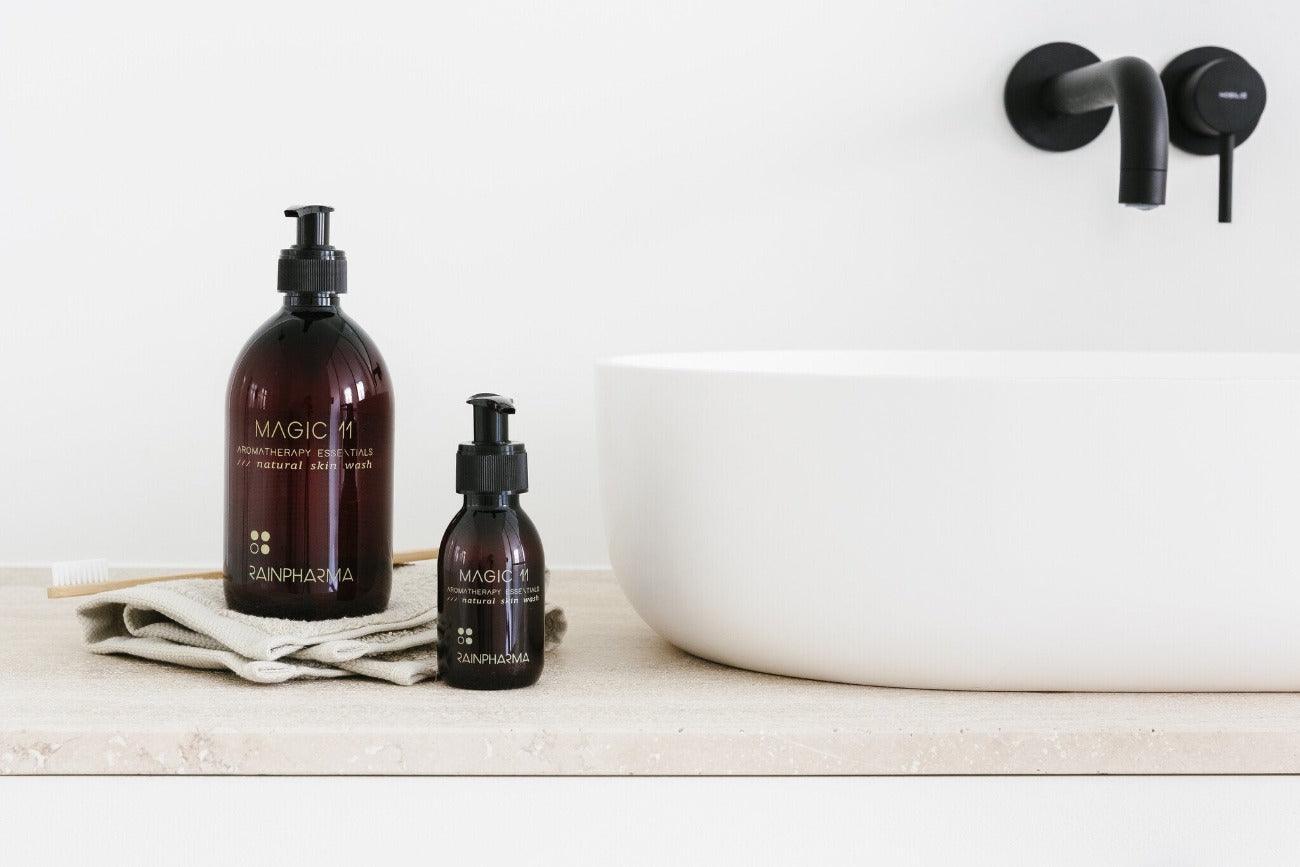 Twee donkere pompflacons met het opschrift "Skin Wash Magic 11" van RainPharma, gemaakt met natuurlijke oliën, staan op een aanrecht in de badkamer naast een witte handdoek, naast een moderne witte wasbak en zwarte kraan.