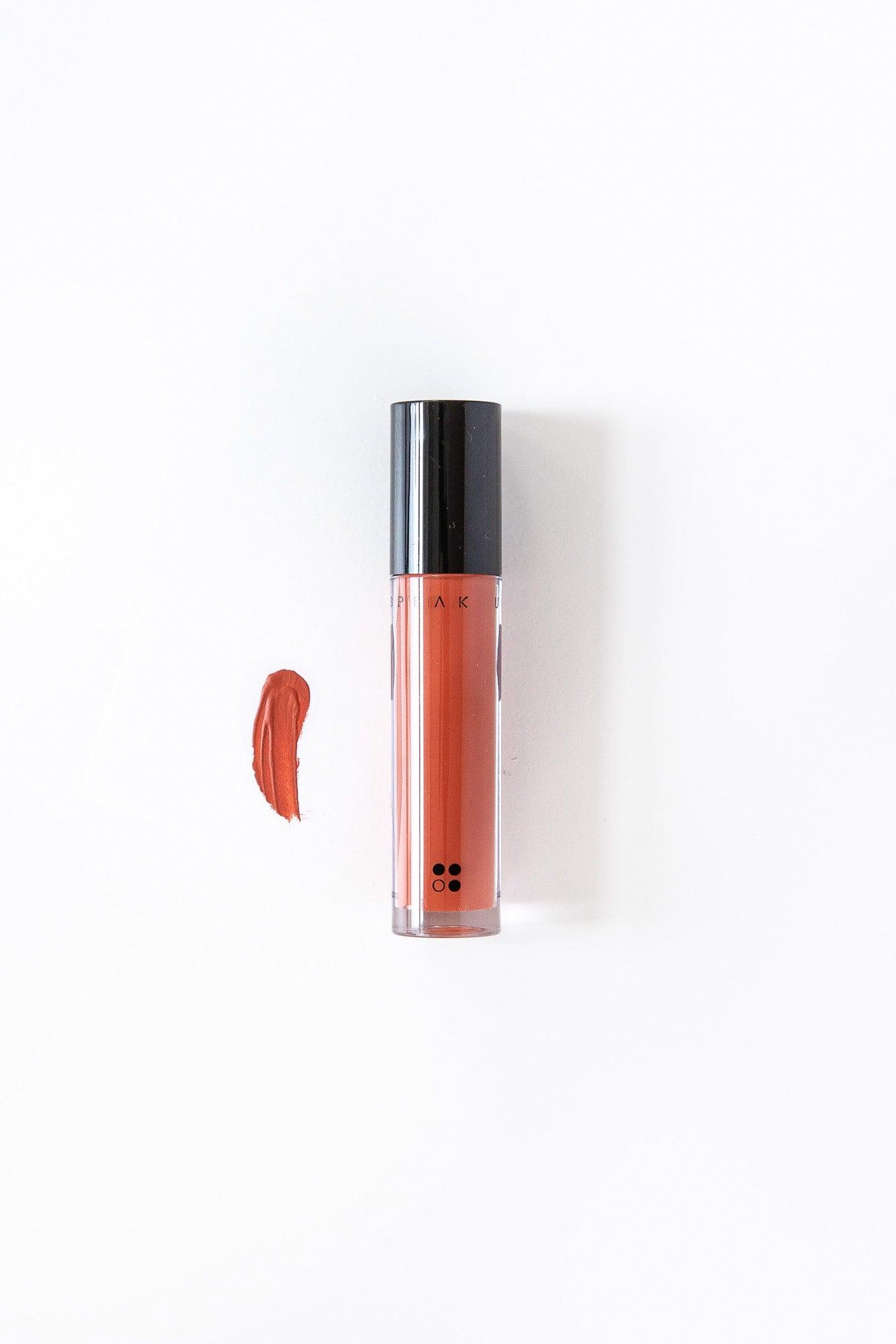 Een tube Speak Up - Natural Lip Tint van RainPharma staat rechtop op een wit oppervlak, met een swatch van dezelfde levendige koraaltint ernaast.