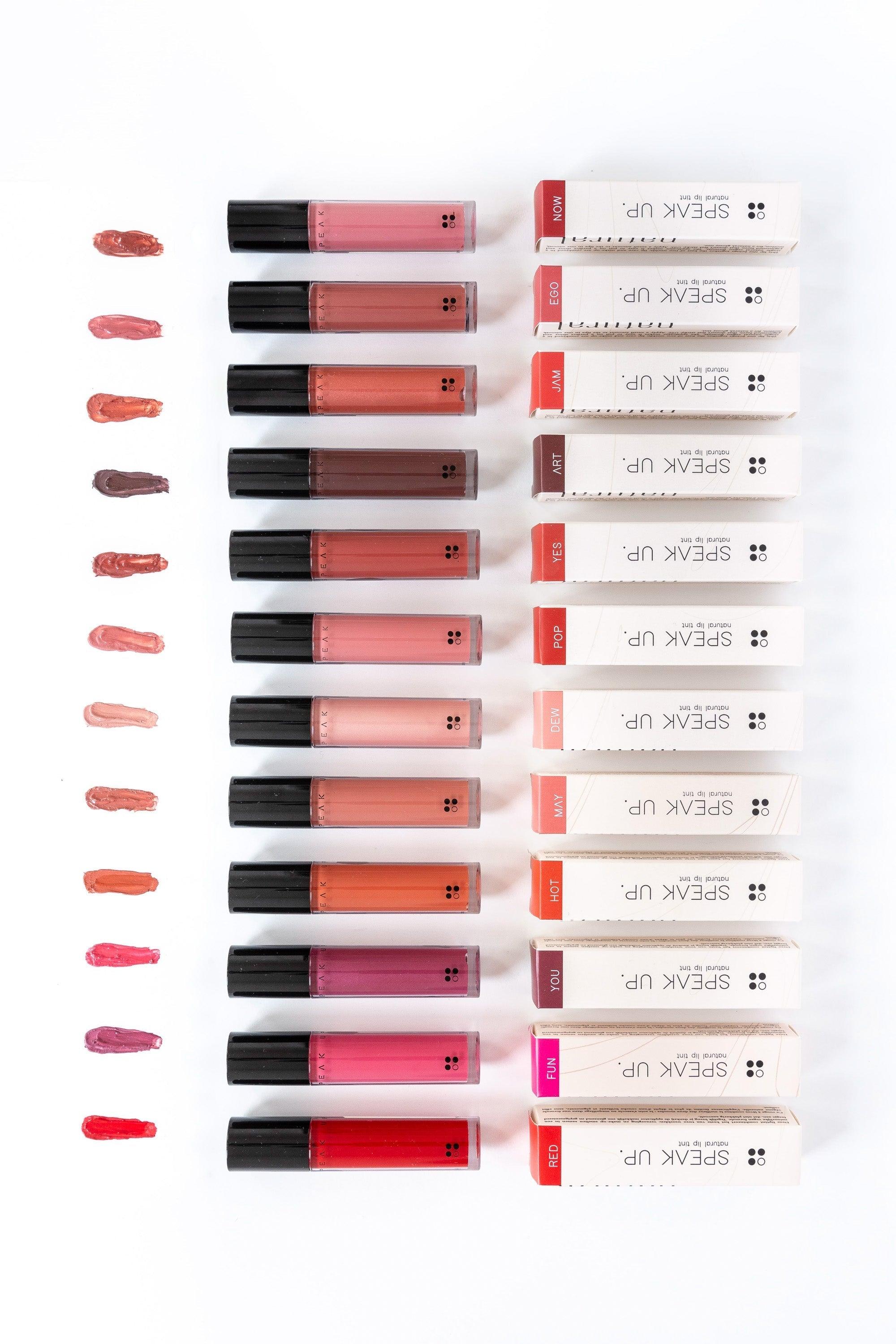 Twaalf tubes van de RainPharma Speak Up - Natural Lip Tint, elk met levendige tinten, worden getoond met swatches. Deze vegan lip tint heeft een glanzend gepigmenteerd finish, en de dozen zijn netjes gestapeld aan de rechterkant.