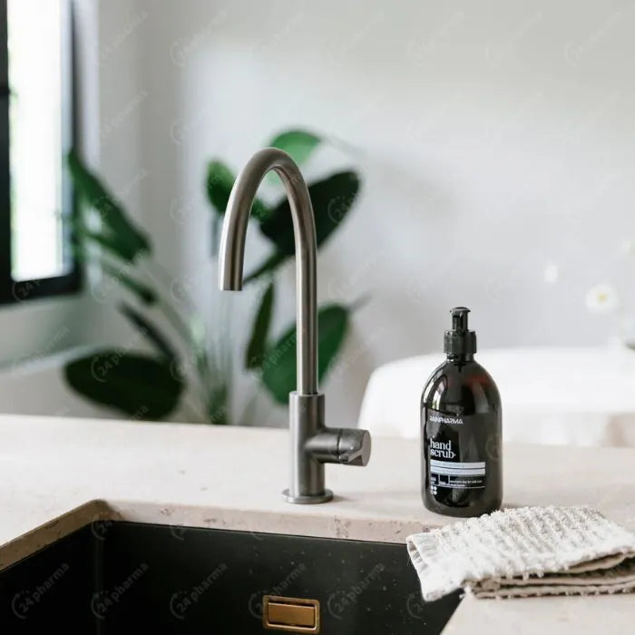 Een modern aanrecht met een gebogen metalen kraan, een 500 ml RainPharma Hand Scrub fles en een textieldoek op het aanrecht; op de achtergrond zijn planten te zien.