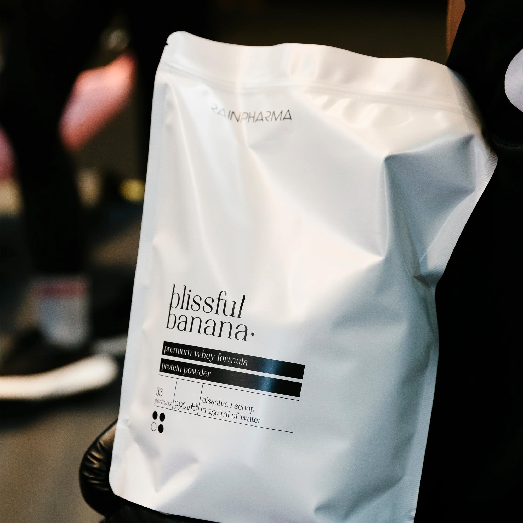 Close-up van een witte Rainpharma zak met het label "XL Shake Blissful Banana - 33 porties" premium whey formule proteïne poeder. De shake biedt een eiwitrijk tussendoortje in bananensmaak met bereidingsinstructies en netto gewicht details op de verpakking.