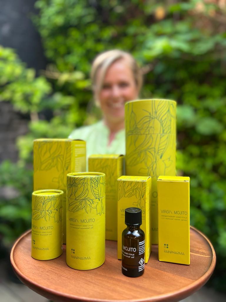 De "Skin Wash - Virgin Mojito" van RainPharma, gemaakt met natuurlijke ingrediënten, is elegant gepresenteerd op een rond houten dienblad, terwijl een persoon glimlacht tegen de weelderige groene achtergrond.
