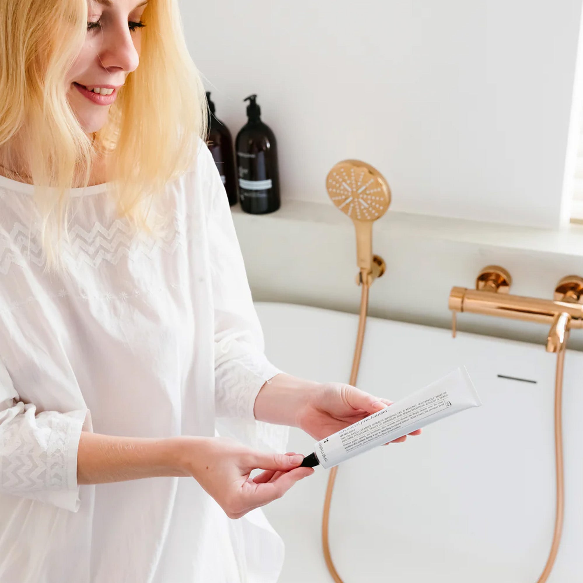 Een vrouw in een witte blouse staat in een badkamer bij een badkuip met goudkleurige accenten, terwijl ze een tube RainPharma Oil Cleanser (Advanced Precleanser) - gelaatsreiniger op oliebasis - vasthoudt en leest.