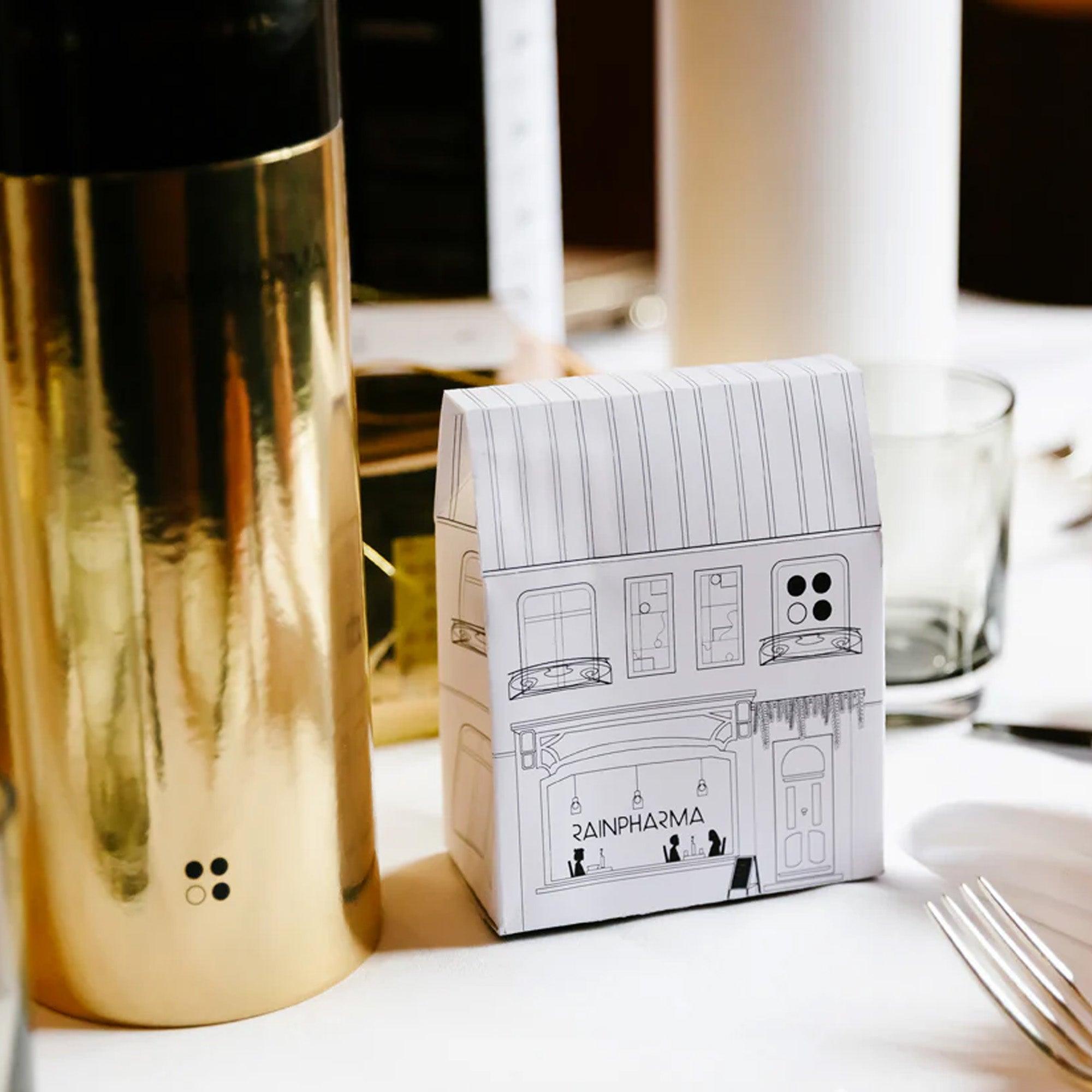 Een decoratief kartonnen huismodel met het label "RAINPHARMA" vult een gouden cilindervormig object aan, wat een spa-gevoel oproept. Deze elegante display past prachtig bij de RainPharma Bistro - Budget Gift set met Skin Wash Lemongrass & After Oil 60 ml, wat het een prachtig cadeau-idee maakt.