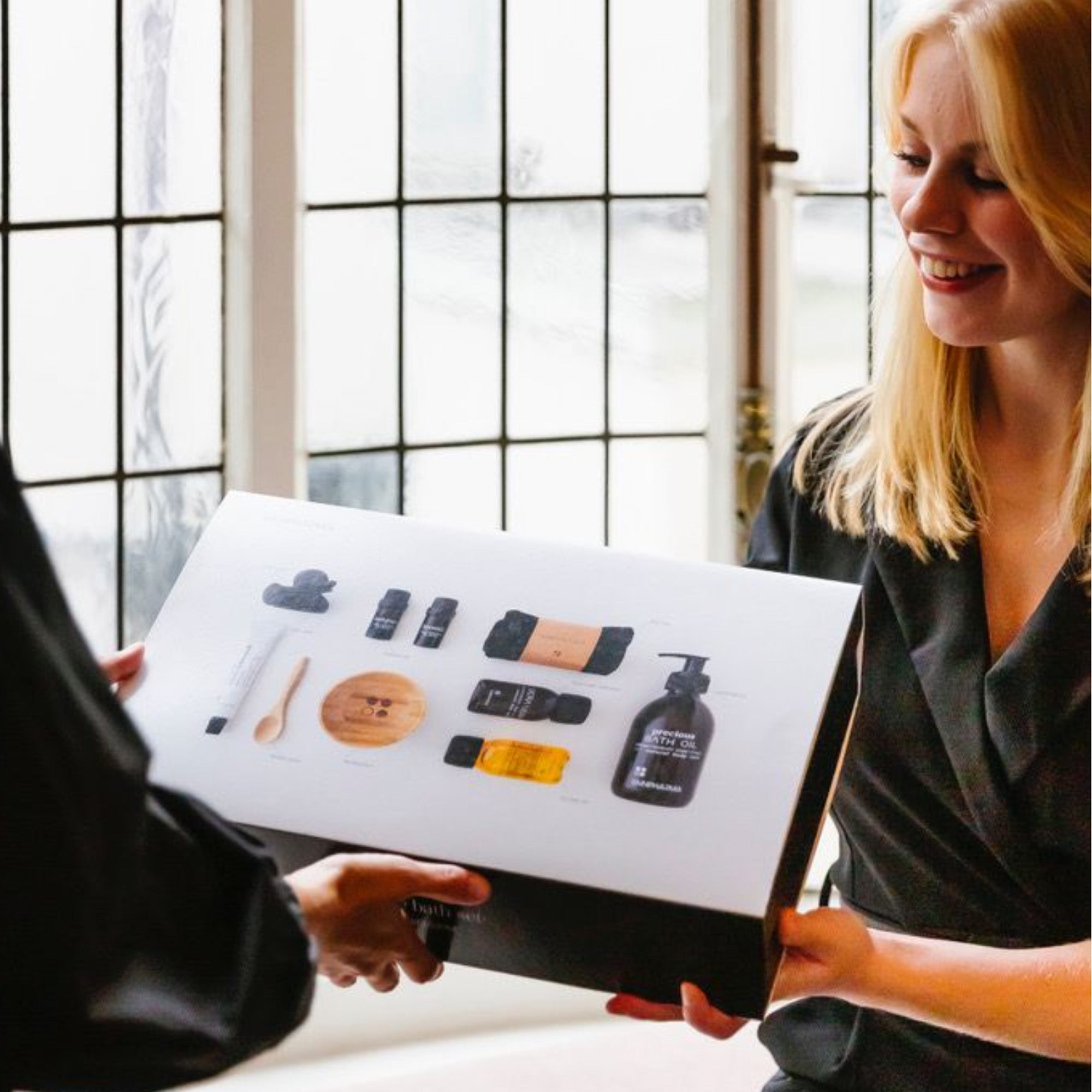 Twee mensen ruilen een samengestelde Luxe Bad Set gevuld met RainPharma huidverzorgings- en wellnessproducten, inclusief badschuim, voor de ultieme spa-ervaring thuis, bij een raam.