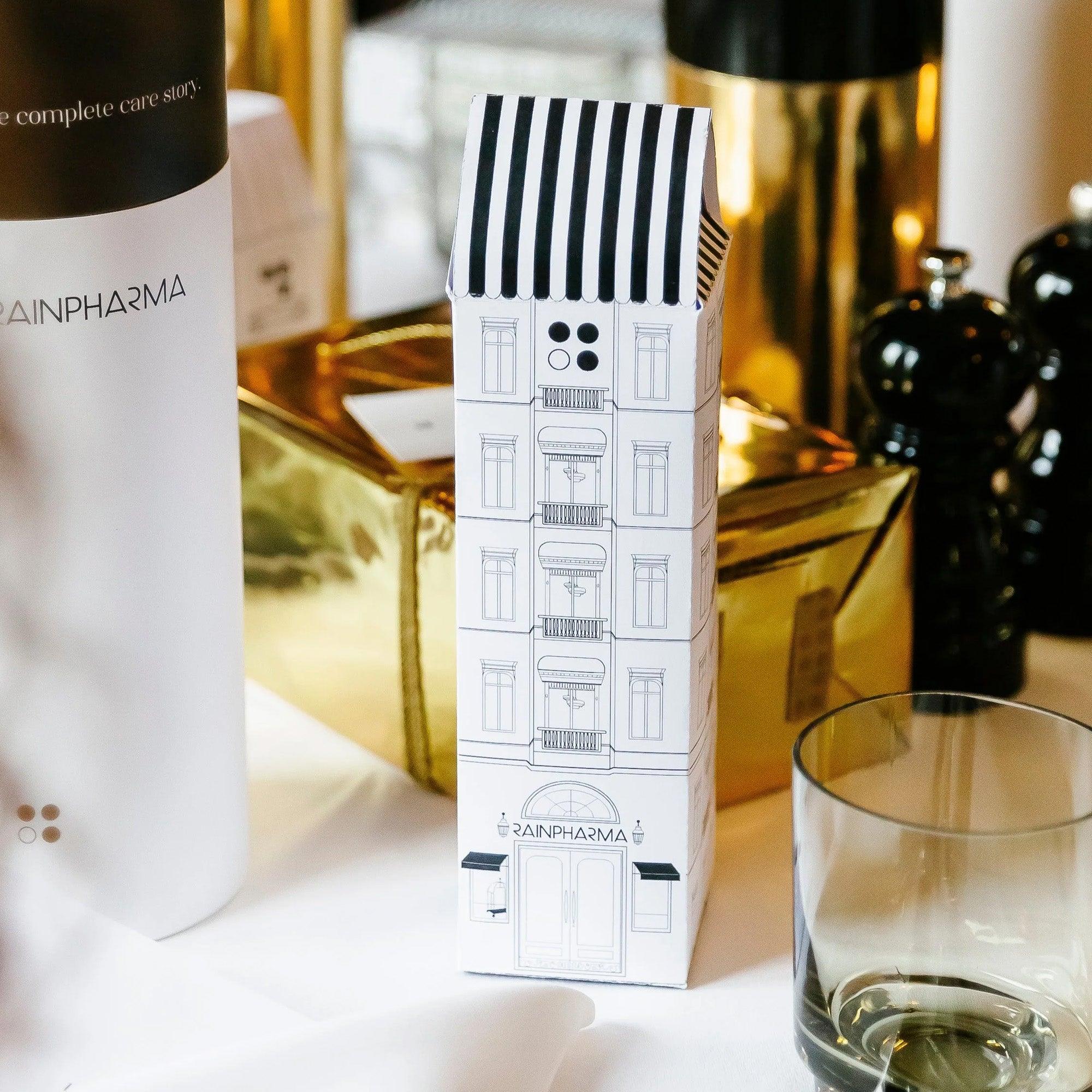 Een decoratieve doos in de vorm van een gebouw staat op een tafel met een glazen, in goud verpakte verpakking en verschillende flessen, waaronder de RainPharma Hotel - Budget Gift set met Skin Wash Lemongrass en Sacred Seven Body Repair Oil 60 ml, perfect om elke ruimte elegantie te geven.