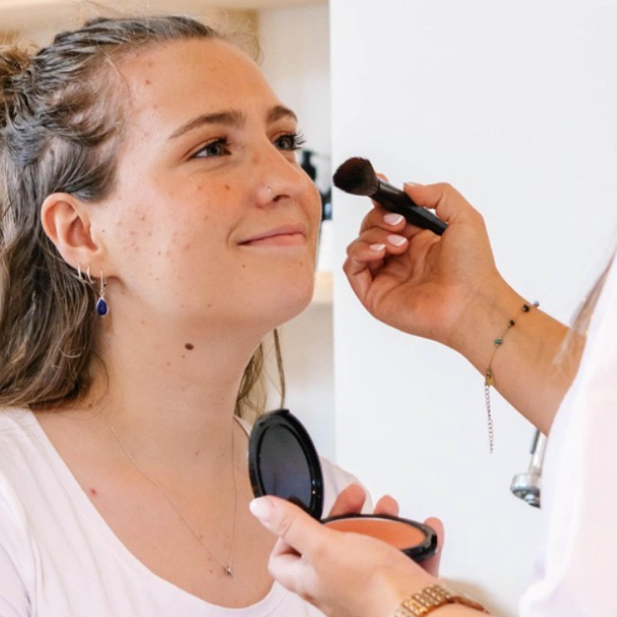 Een persoon brengt Puur Living make-up aan op het gezicht van een lachende vrouw terwijl ze een compact spiegeltje vasthoudt - perfect om dit prachtige Cadeaubon Make-up verwenmoment voor twee vast te leggen.