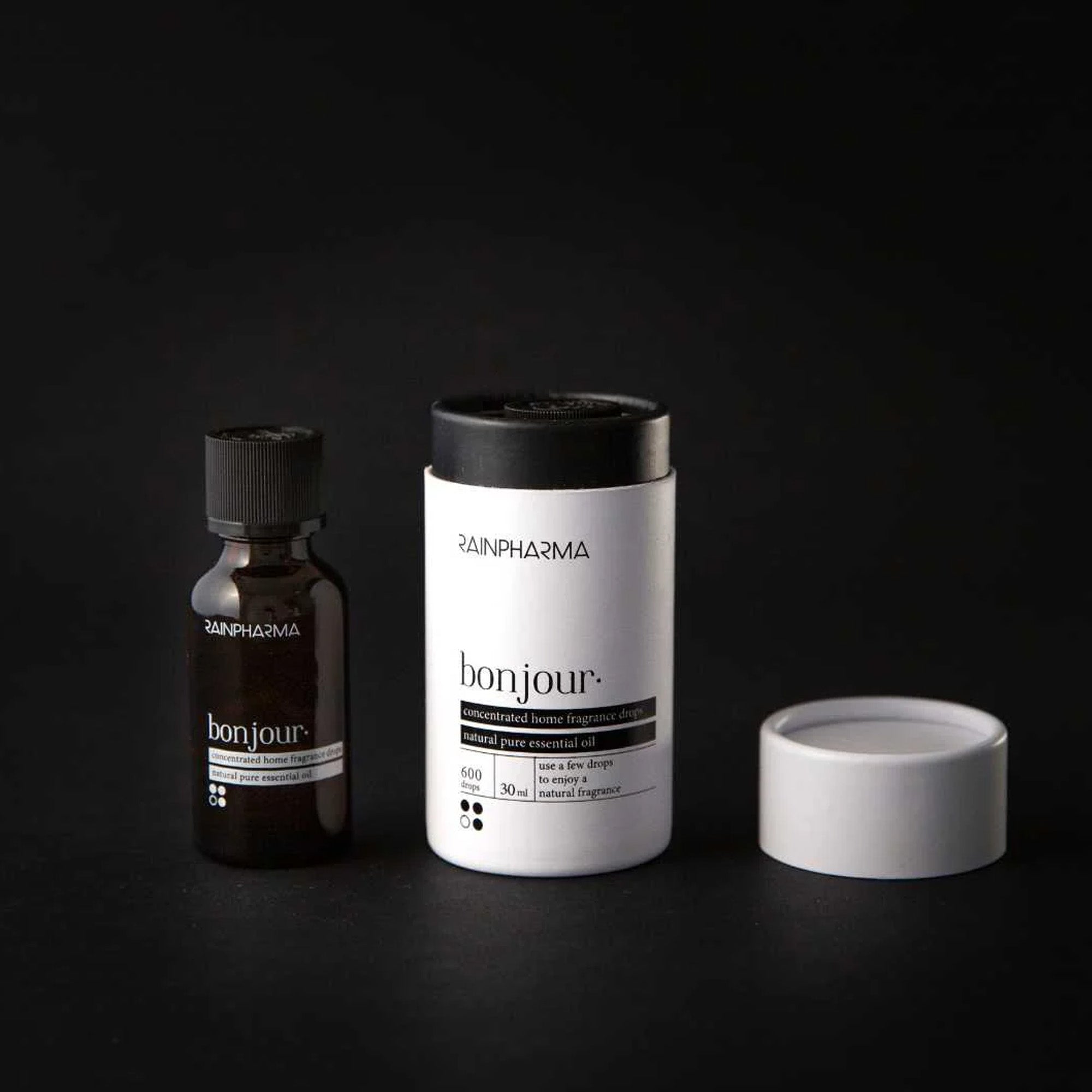 Twee RainPharma-artikelen staan op een donkere achtergrond: een klein bruin flesje en de witte cilindrische Essential Oil - Bonjour 30ml-container, met het deksel opzij. Beroemd om premiumkwaliteit.