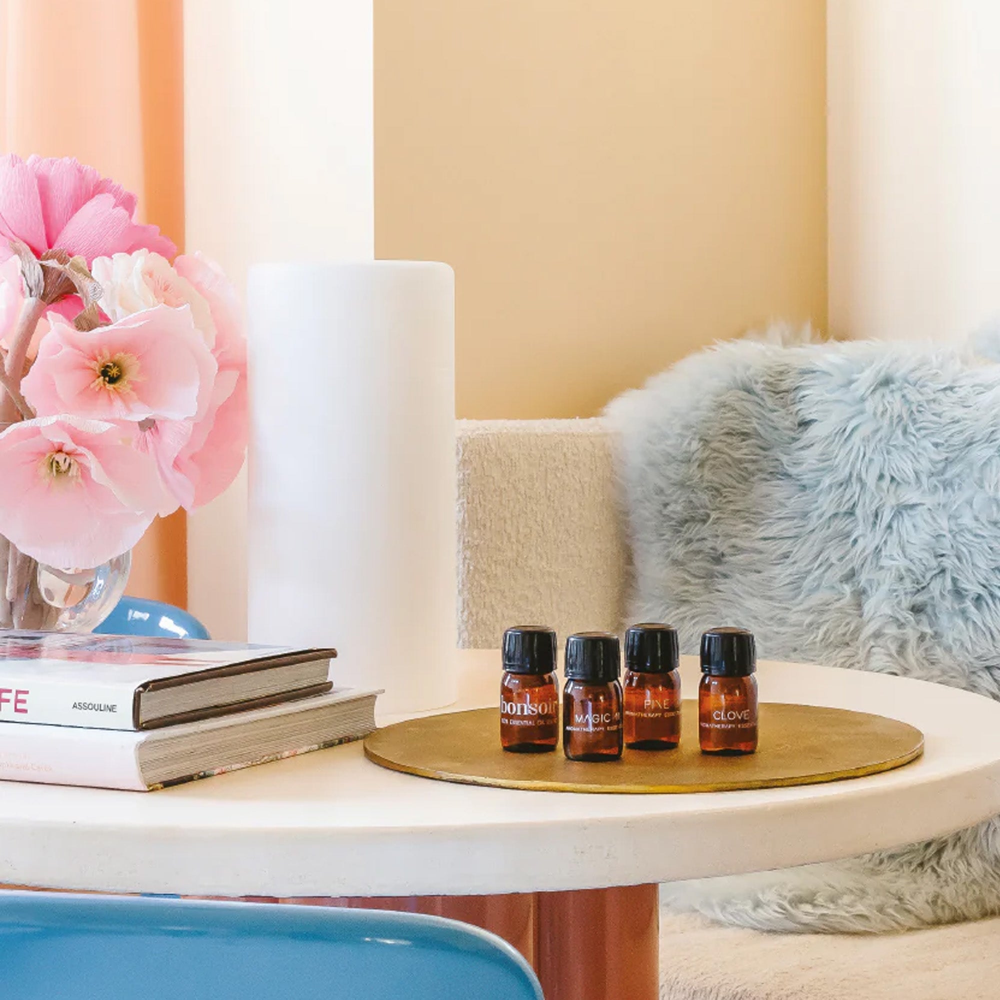 Op een ronde tafel staat een dienblad met vier flesjes "Essentiële Olie - Bonjour 30ml" van RainPharma, naast een vaas met roze bloemen, wat boeken en een zacht kussen op de achtergrond.