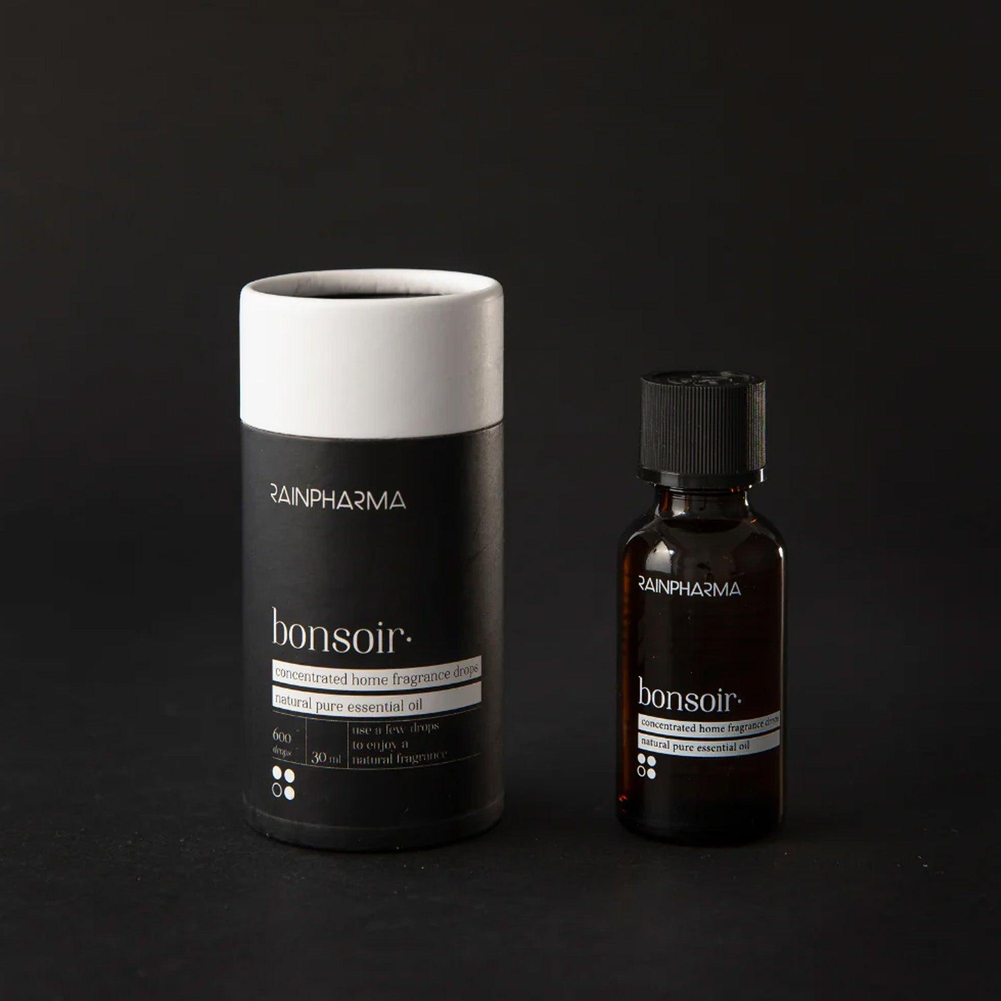 Een klein bruin flesje met het opschrift "Essentiële Olie - Bonsoir 30 ml" van RainPharma staat naast een cilindervormige zwart-witte verpakking. Beide staan elegant op een strakke zwarte achtergrond en belichamen de essentie van natuurlijke producten.
