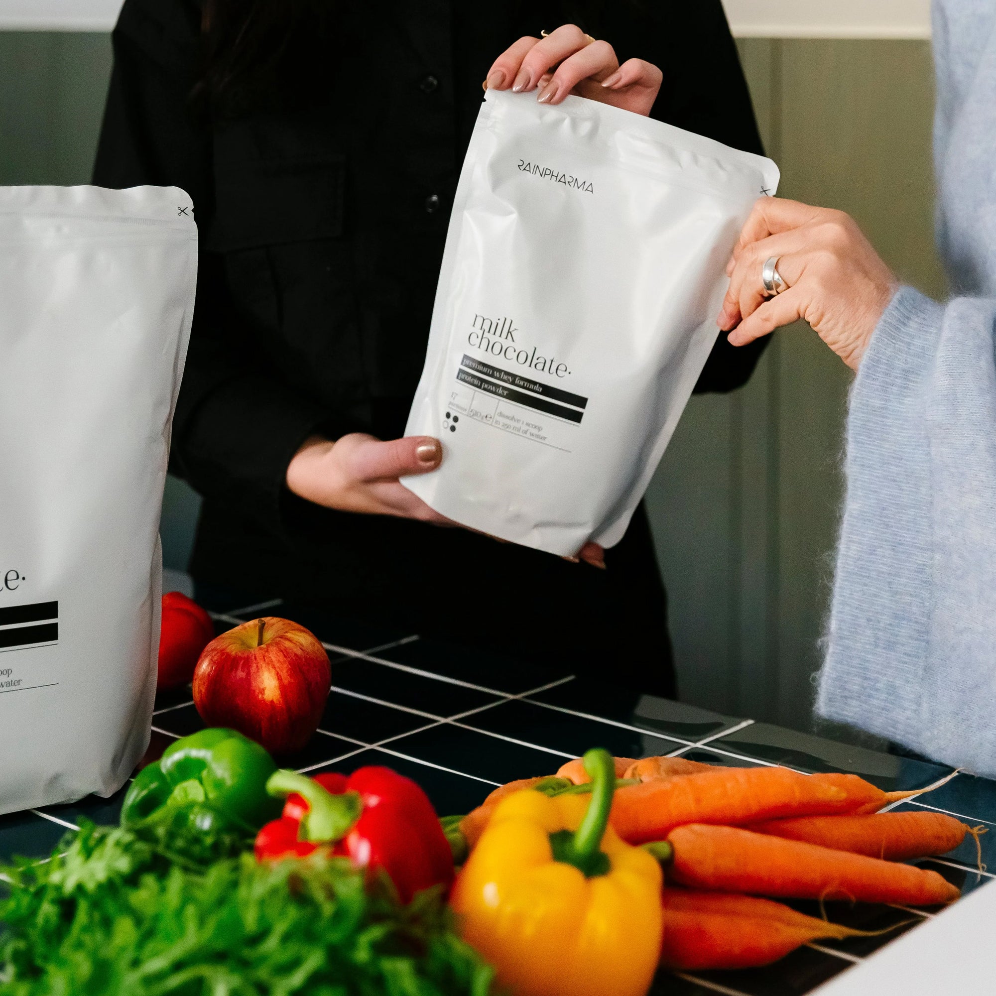 Twee mensen met witte RainPharma Nutrition Program zakjes bij een tafel met appels, wortels, paprika&