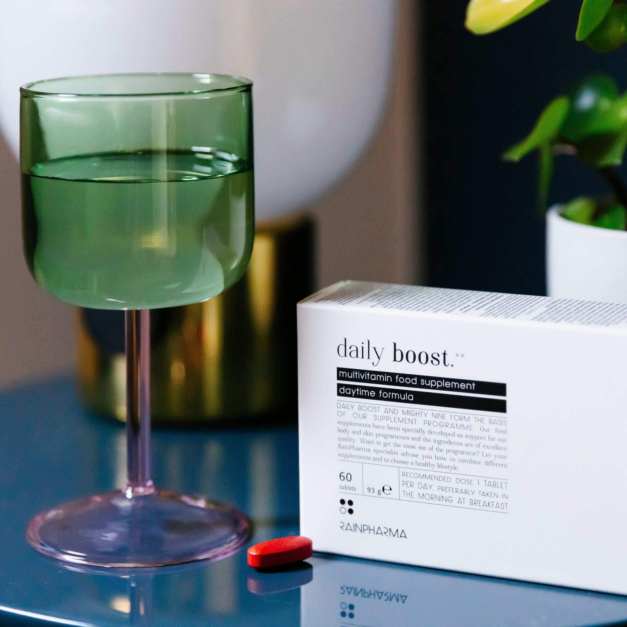 Een groen glas water, een doos met het RainPharma Nutrition Program multivitaminesupplement en een rode tablet rusten op een reflecterend blauw oppervlak en tonen het RainPharma Nutrition Program.