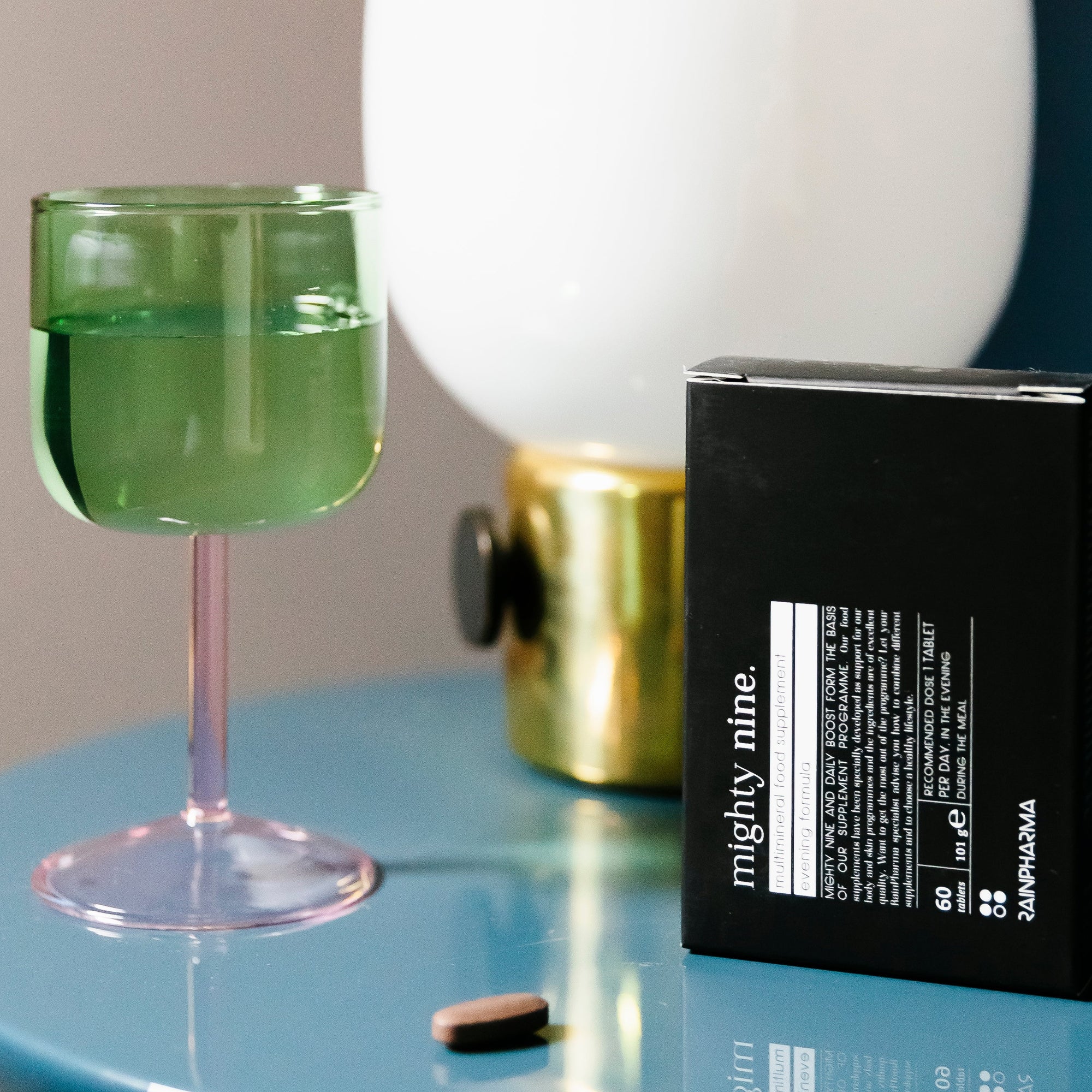 Een groen drankje in een glas, een RainPharma Nutrition Program doos, een supplementtablet en een lamp op een glanzend blauw tafeltje-ideale benodigdheden voor je dagelijkse RainPharma Nutrition Program routine.