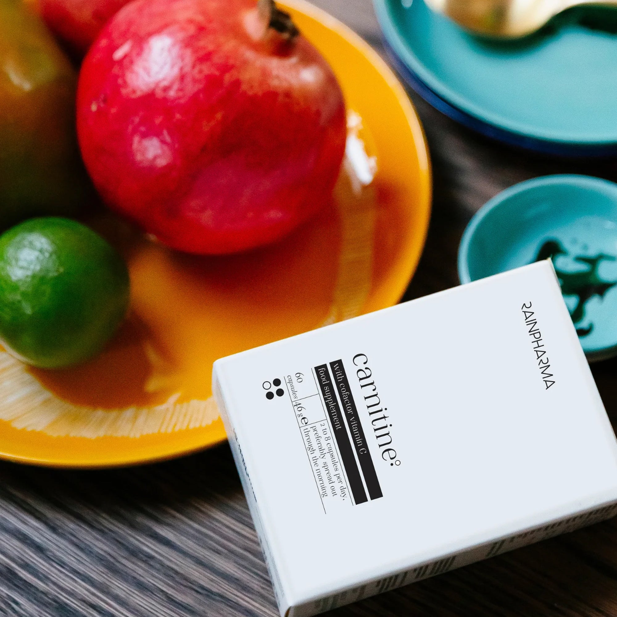 Een witte doos met het label "RainPharma" van het RainPharma Nutrition Program staat op een tafel naast een geel bord met een granaatappel, limoen en groene appel - een uitnodigend tafereel dat een bewuste keuze in voeding en voedingssupplementen suggereert.