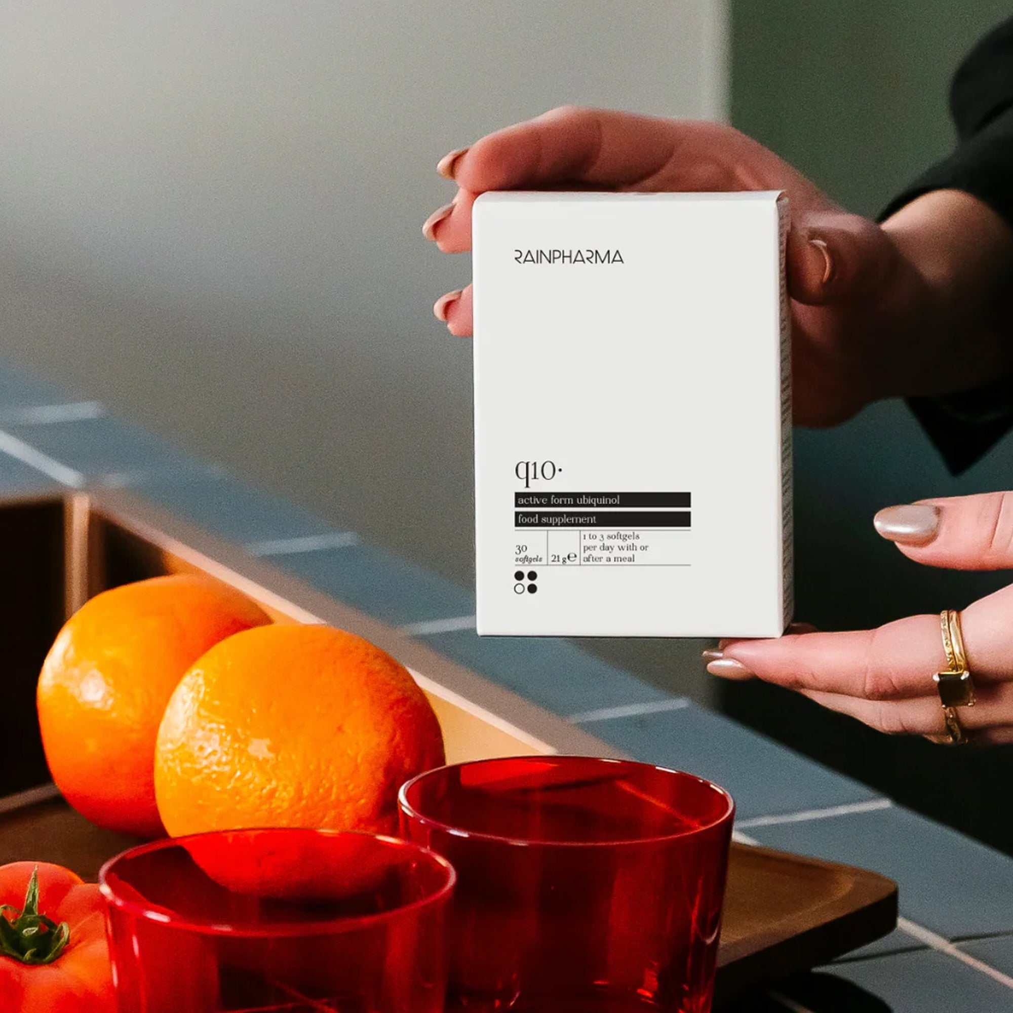 Iemand houdt een witte doos van het RainPharma Nutrition Program vast, een kwaliteitssupplement van RainPharma, naast een toonbank met sinaasappels, een tomaat en rode glazen.