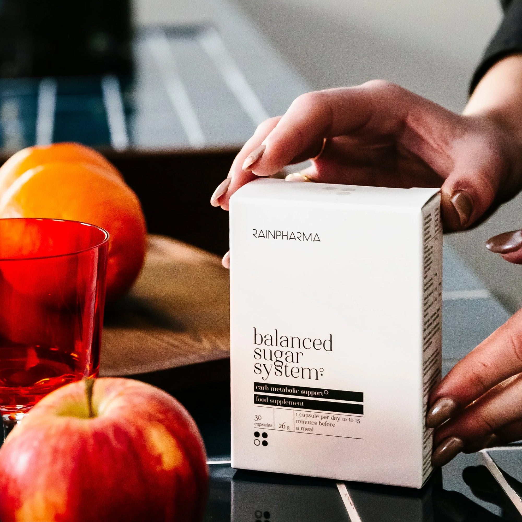 Een hand houdt een doos vast met het label "Puur Living Restart Programma" van RainPharma, onderdeel van hun Smart Nutrition Project. Ernaast liggen rode appels, een rood glas en een houten dienblad op een reflecterend zwart oppervlak.