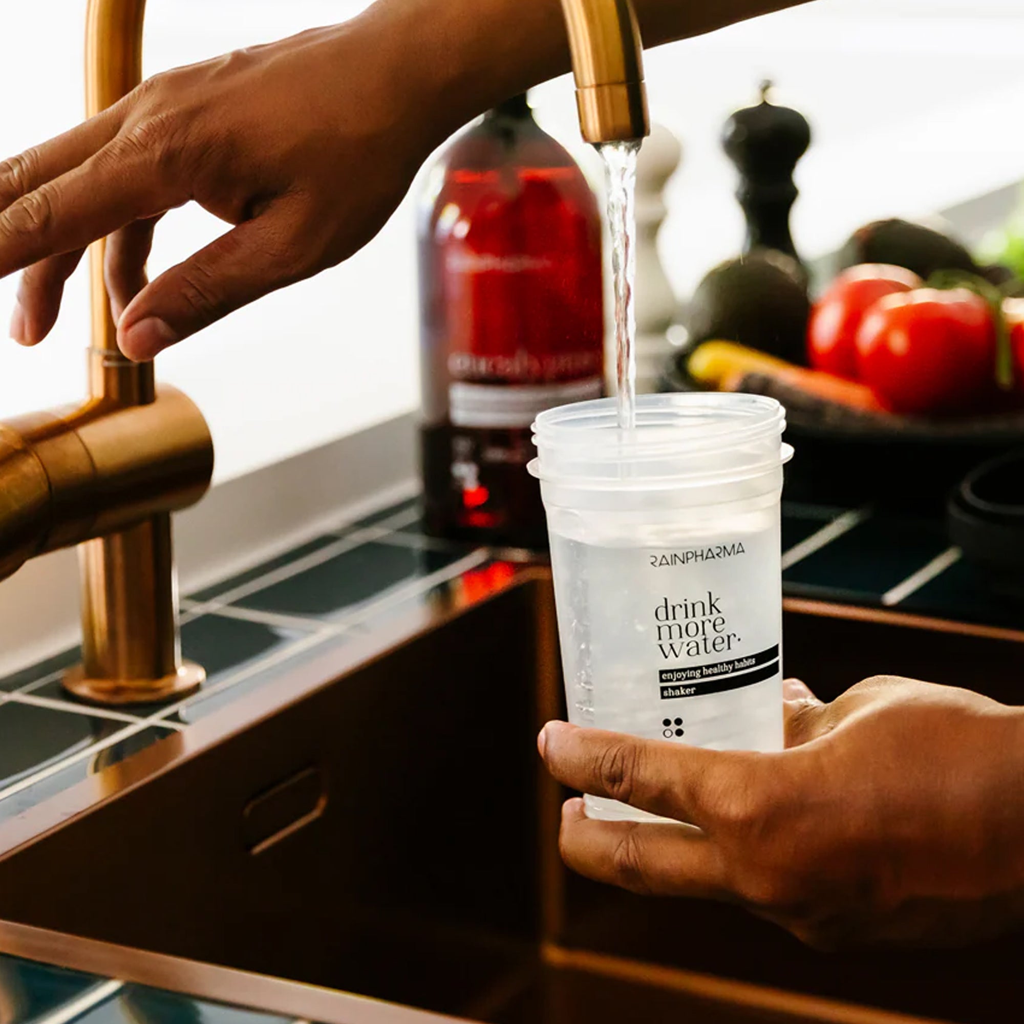 Een persoon vult een doorzichtige plastic beker met de tekst "drink meer water" met water uit een keukenkraan, terwijl op het aanrecht op de achtergrond de supplementen van RainPharma Nutrition Program en andere producten zichtbaar zijn.