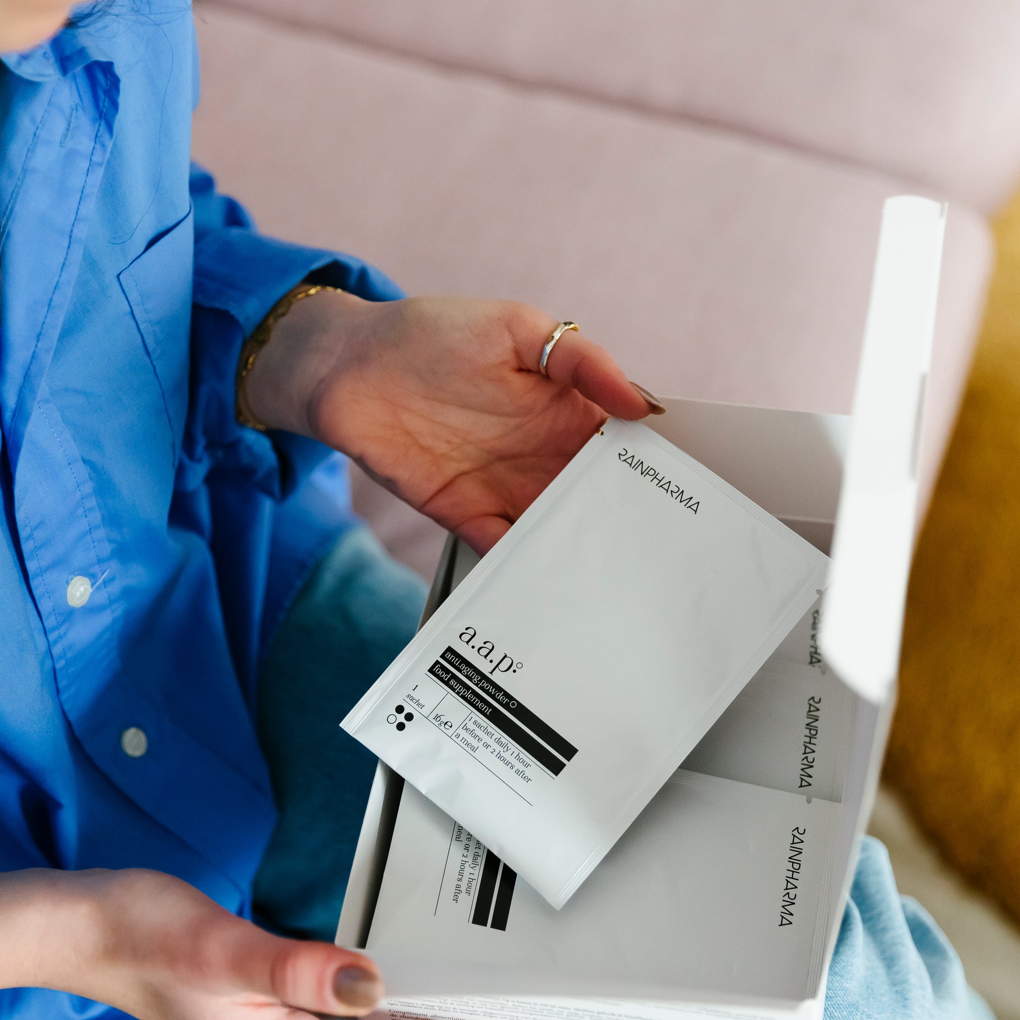 Een persoon in een blauw shirt houdt zittend een open witte doos met verschillende witte zakjes vast met de tekst "RainPharma Collageen Supplement - AAP (anti.aging.powder)" van RainPharma.