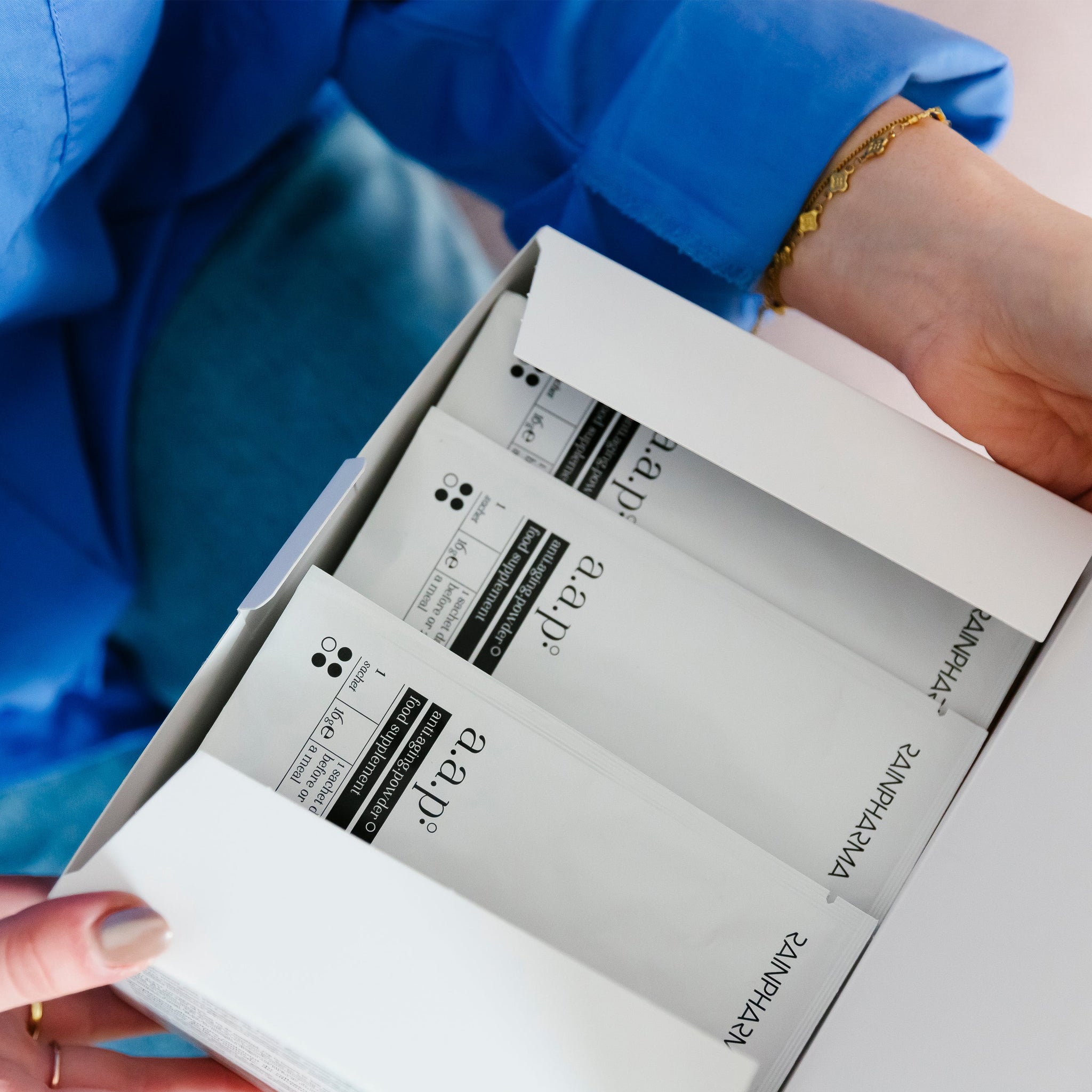 Een persoon opent een witte doos RainPharma Collageen Supplement - AAP (anti.aging.powder), waarop verschillende pakjes staan met termen als "aanpassen", "AM/PM", ingrediënten zoals glucosamine en zichtbare pictogrammen op de verpakking.