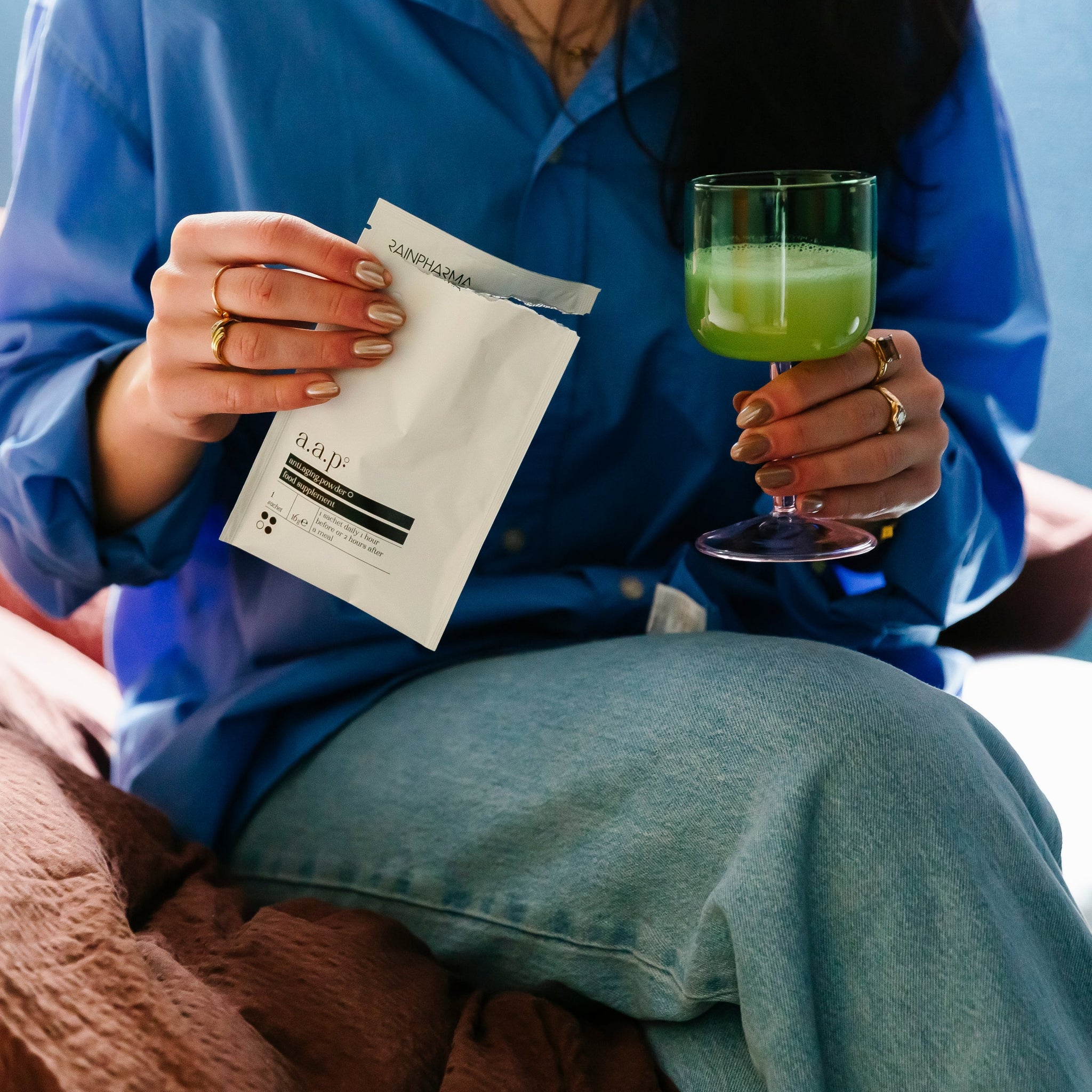 Een persoon in een blauw shirt houdt een geopend RainPharma Collageen Supplement - AAP (anti.aging.powder) pakje in de ene hand en een groen drankje in een glas met steel in de andere.