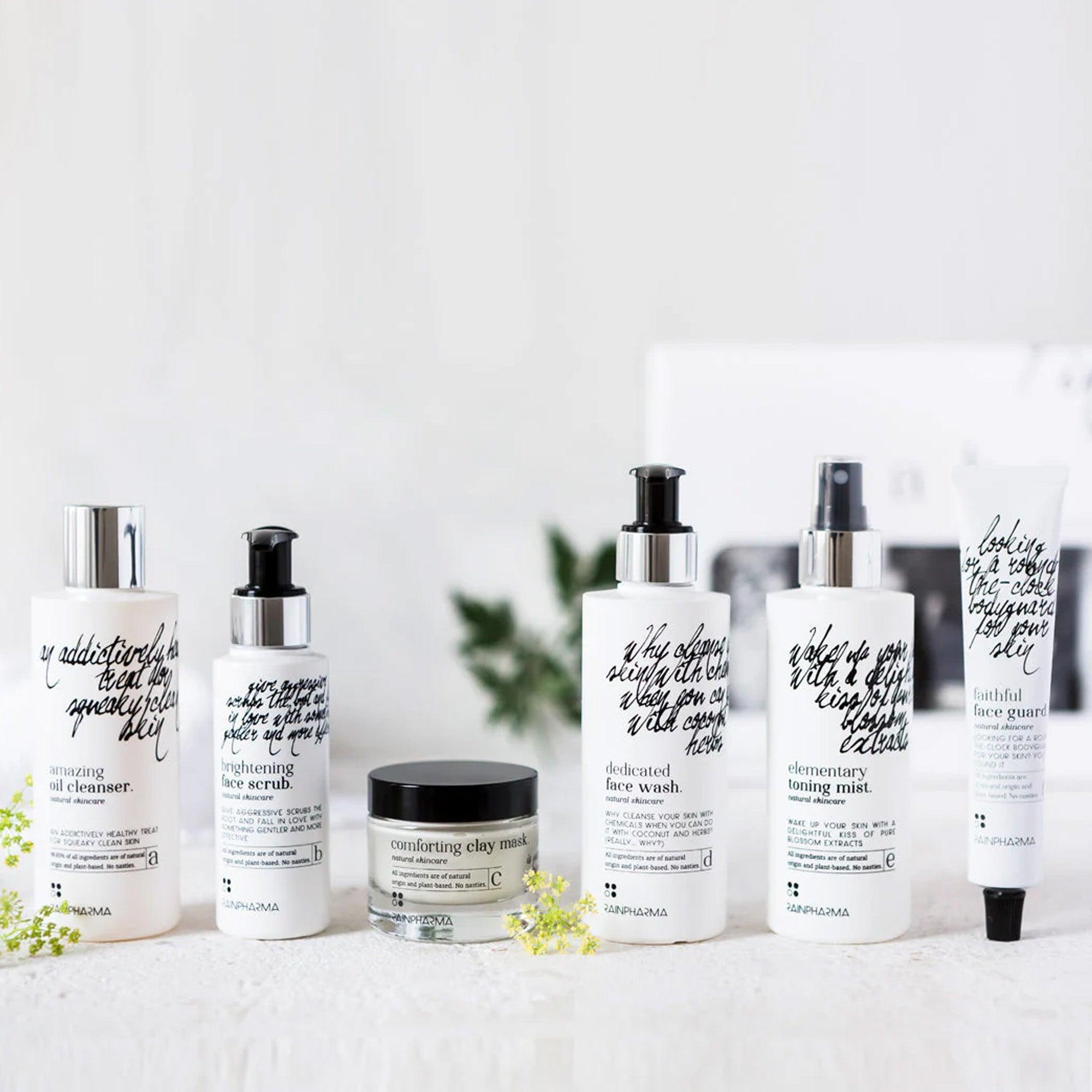 De RainPharma Complete Skincare set (abcdef promobox 4+2 GRATIS) toont zes natuurlijke huidverzorgingsproducten in zwarte tekst op witte flessen en tubes, netjes gerangschikt. De collectie omvat een doorzichtige pot op een licht oppervlak met een wazige achtergrond, wat de holistische benadering benadrukt.