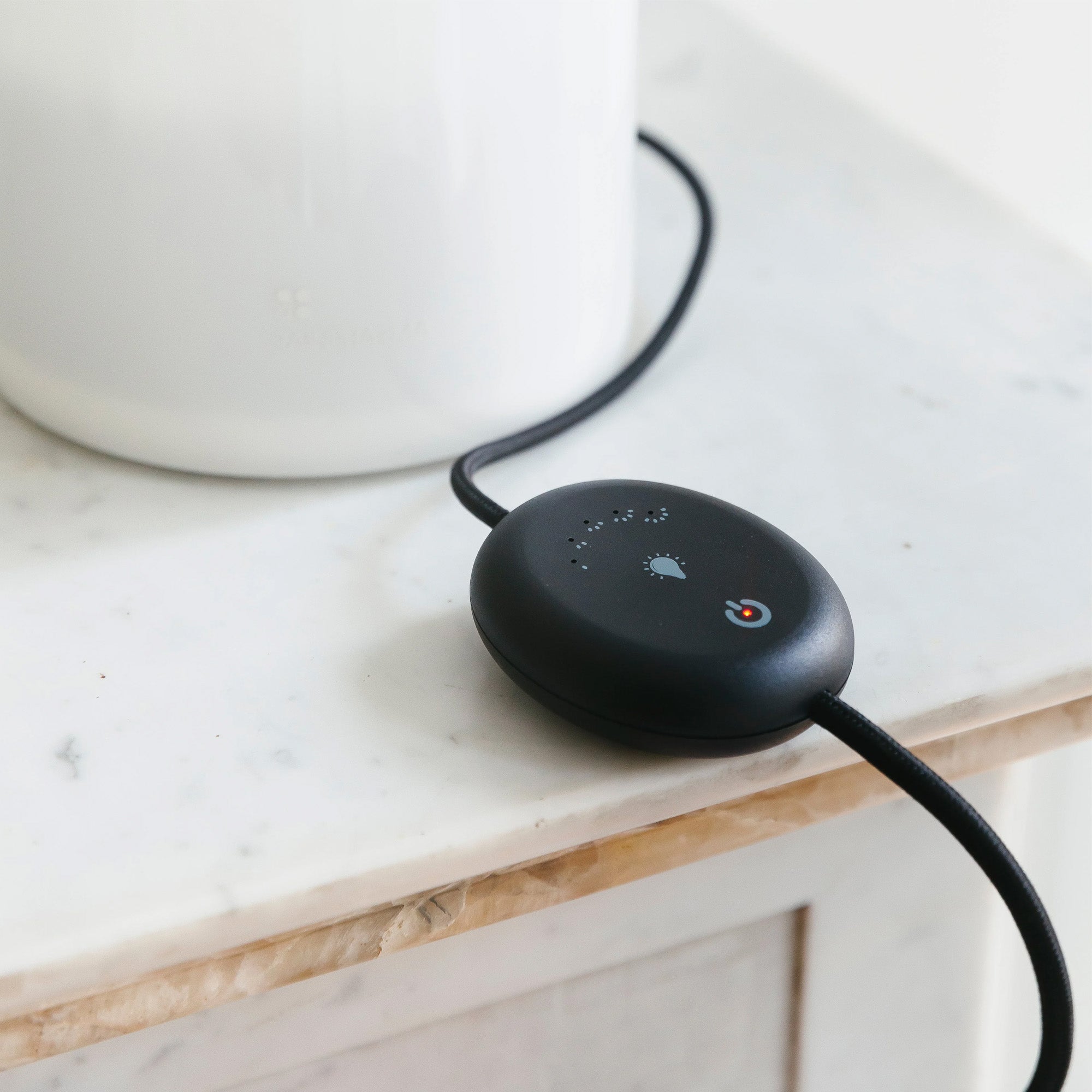 Een strakke, zwarte Aroma Diffuser Set van RainPharma, met de Aurora Base en een zilveren glazen hoes met snoer, staat op een marmeren oppervlak naast een witte houder, wat het moderne design benadrukt.