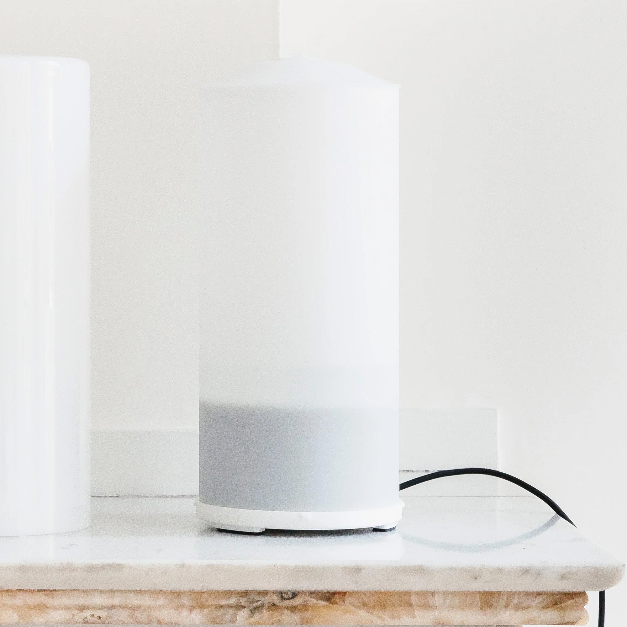 Een RainPharma Aroma Diffuser Set, bestaande uit de Aurora Base en Moonlight Glass Sleeve in een strak cilindervormig ontwerp, rust op een marmeren oppervlak met een zwart snoer dat uit de basis steekt.