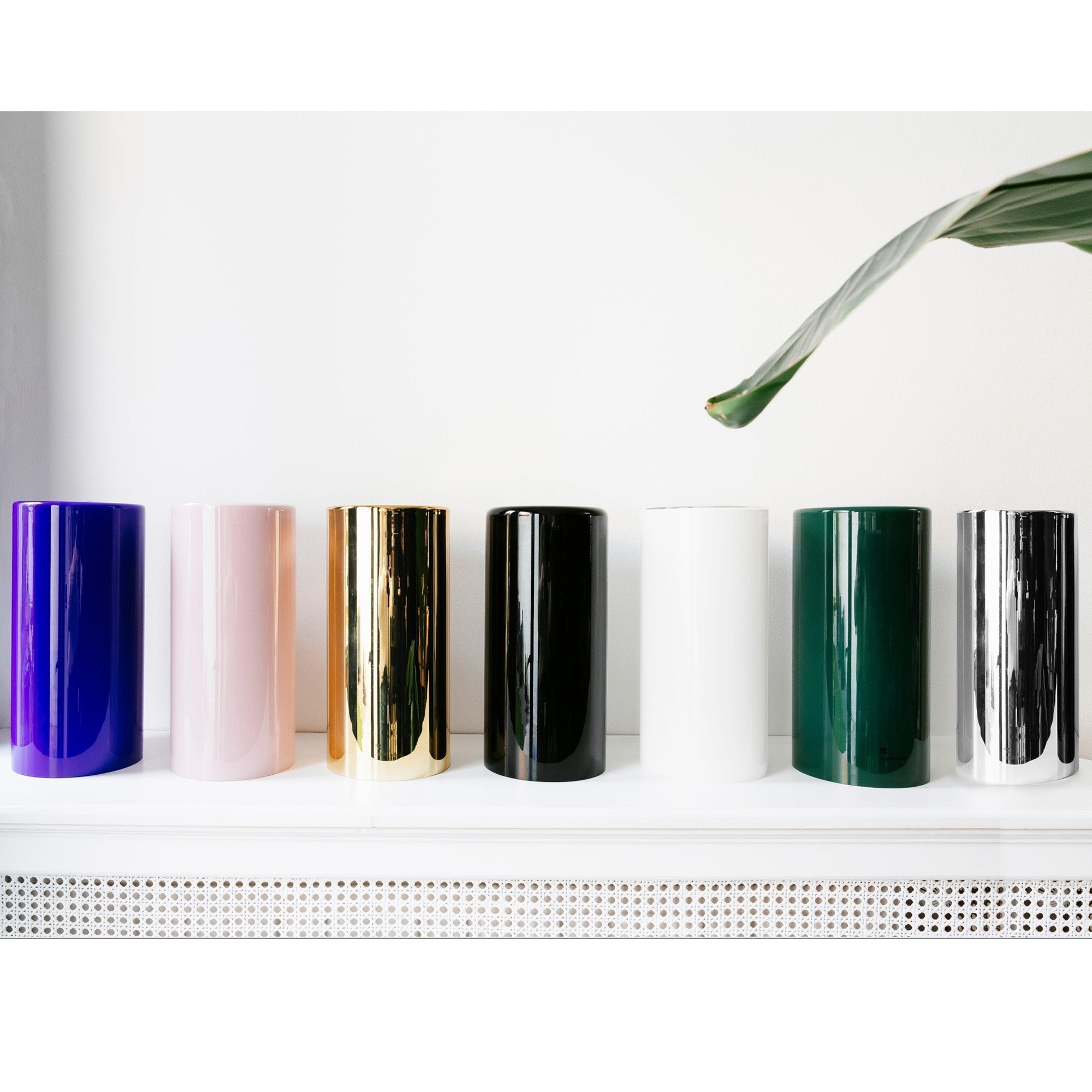 Een rij cilindervormige vazen, vergelijkbaar met de Aroma Diffuser - Aurora Glass Sleeve van RainPharma, worden elegant tentoongesteld op een wit oppervlak in verschillende kleuren: blauw, roze, goud, zwart, wit, groen en zilver.