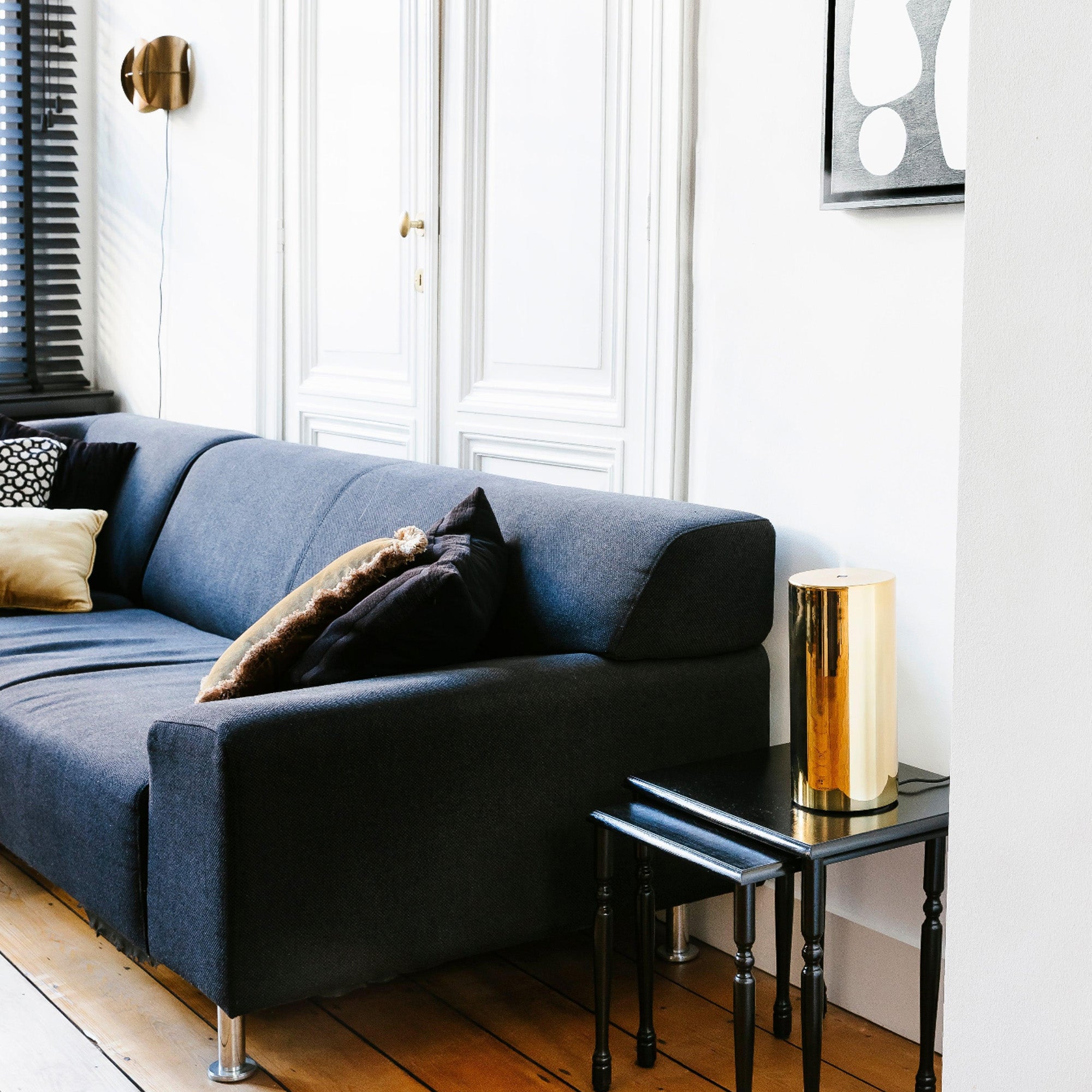 Een moderne woonkamer met een donkergrijze bank met diverse kussens, een RainPharma Aroma Diffuser Set op het zwarte bijzettafeltje, een gouden lamp en abstracte wandkunst, allemaal gerangschikt op een lichte houten vloer.