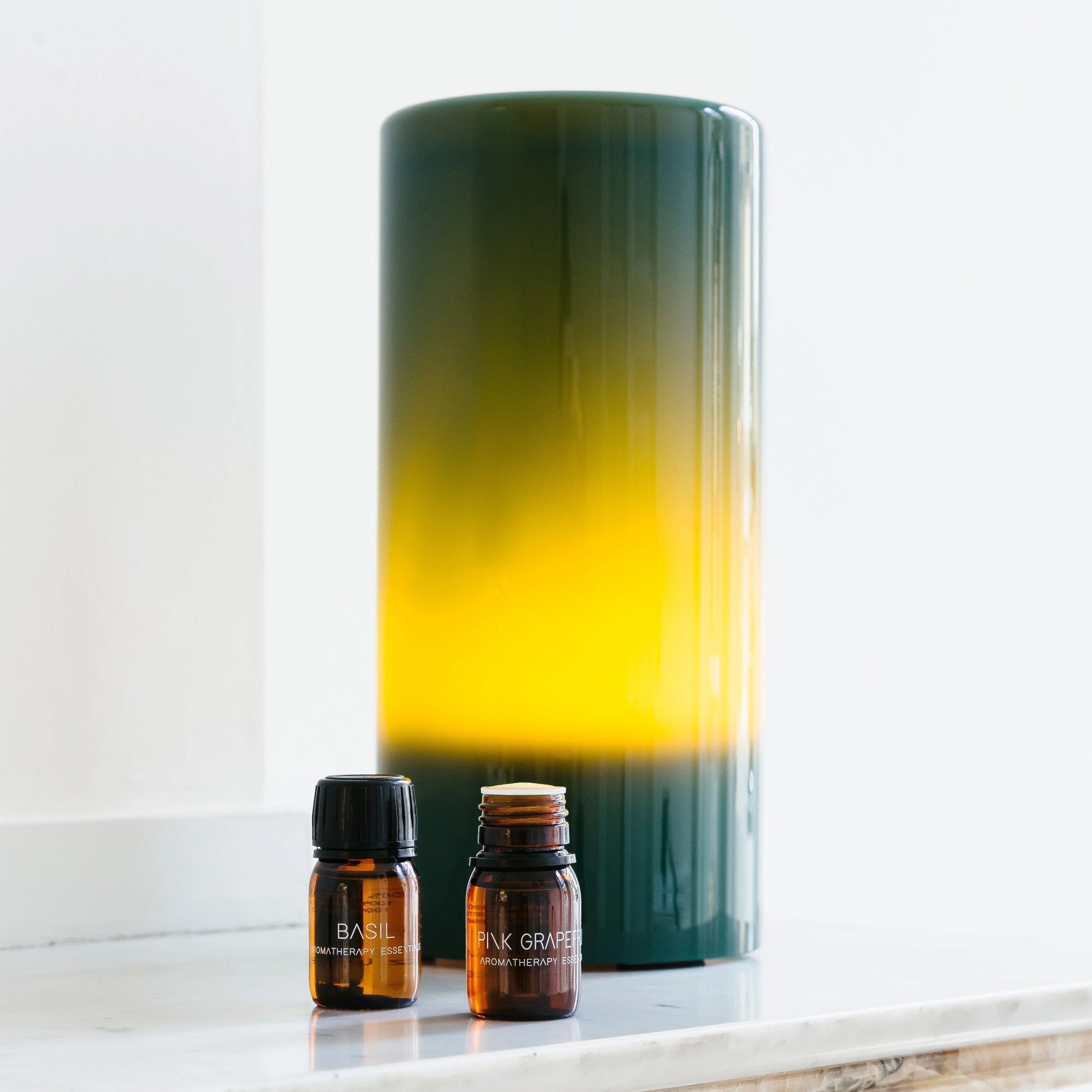 De RainPharma Aroma Diffuser Set - Aurora Base + Glass Sleeve in Dark Forest bestaat uit twee kleine bruine flesjes met de labels "Basil" en "Pink Grapefruit". Deze flesjes staan voor een grote, groen-gele ombre diffuser, die elke ruimte verfraait met zijn geurige mist die is gemaakt met behulp van ultrasone technologie.
