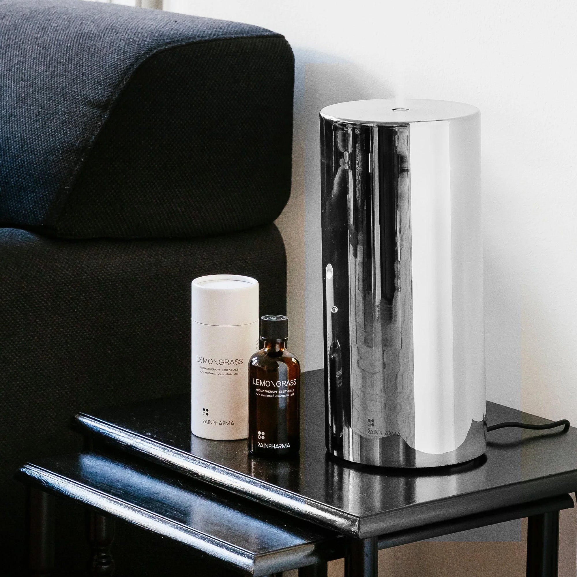 Een strakke metalen luchtreiniger met ultrasone technologie staat op een klein zwart tafeltje naast een zwarte bank, samen met een houder en de RainPharma Aroma Diffuser Set - Aurora Base + Glass Sleeve in zilver, met het label Lemongrass.