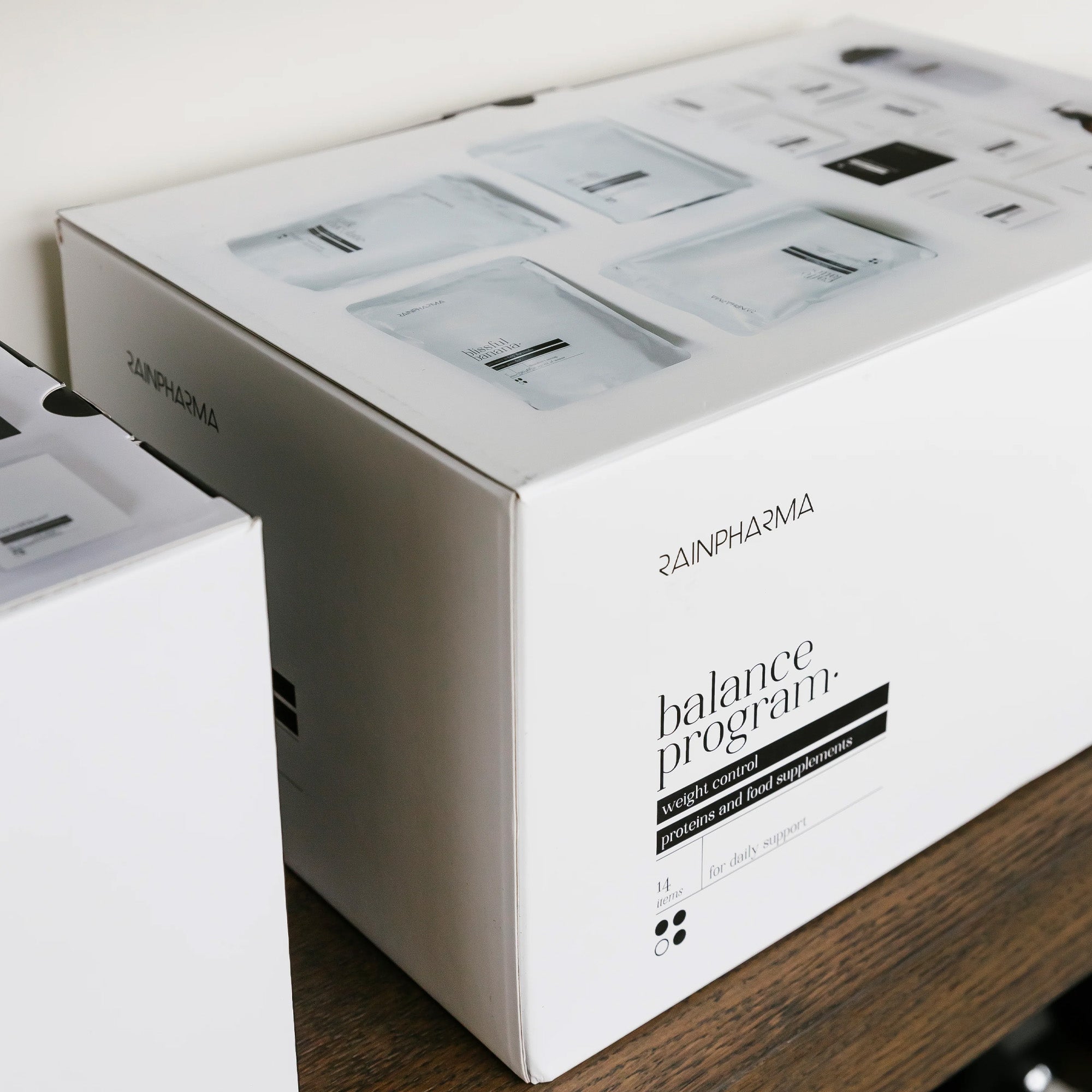 Een wit RainPharma Balance Program (Dieet Programma) doosje staat op een houten oppervlak met productinformatie en afbeeldingen van proteïneshake zakjes - ideaal voor iedereen die kiest voor natuurlijke voeding.