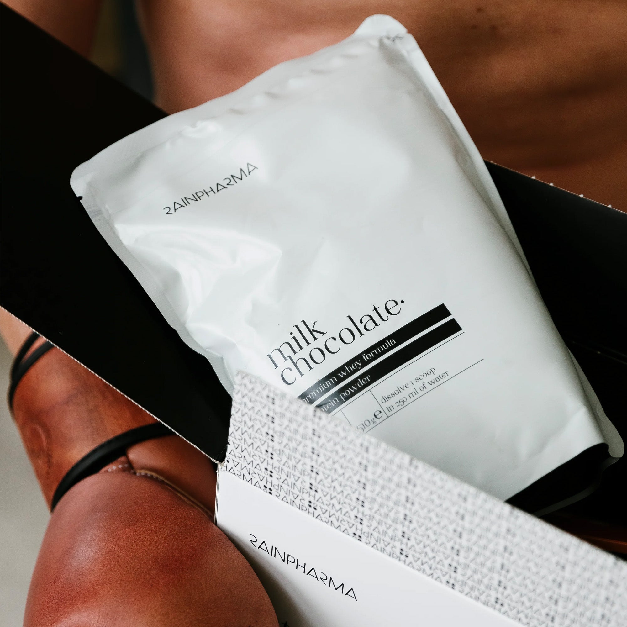 Een witte verpakking met het etiket "melkchocolade" van RainPharma wordt gedeeltelijk verwijderd uit een doos van het RainPharma Balance Program - Dieet Programma; op de achtergrond zijn benen zichtbaar, wat duidt op een natuurlijke dieet- of proteïneroutine.