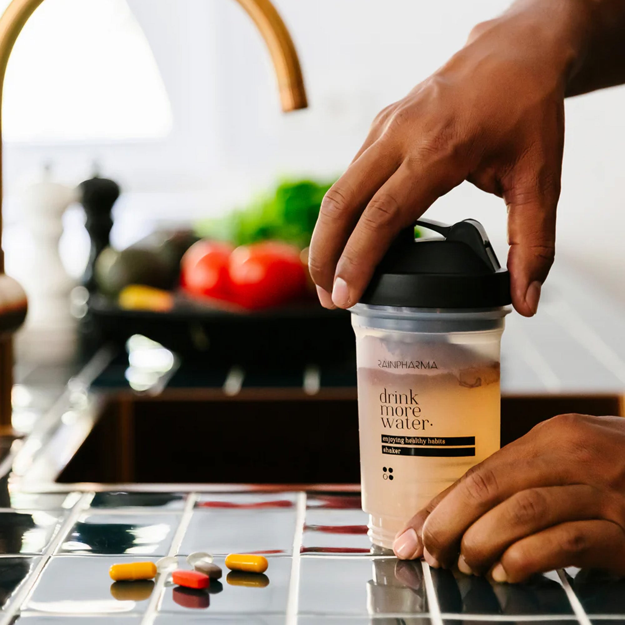 Een hand houdt een shakebeker met "drink meer water" vast bij een gootsteen, terwijl pillen op het aanrecht rusten. Het RainPharma Balans Programma (Dieet Programma) en vage groenten op de achtergrond benadrukken de focus op natuurlijke voeding.