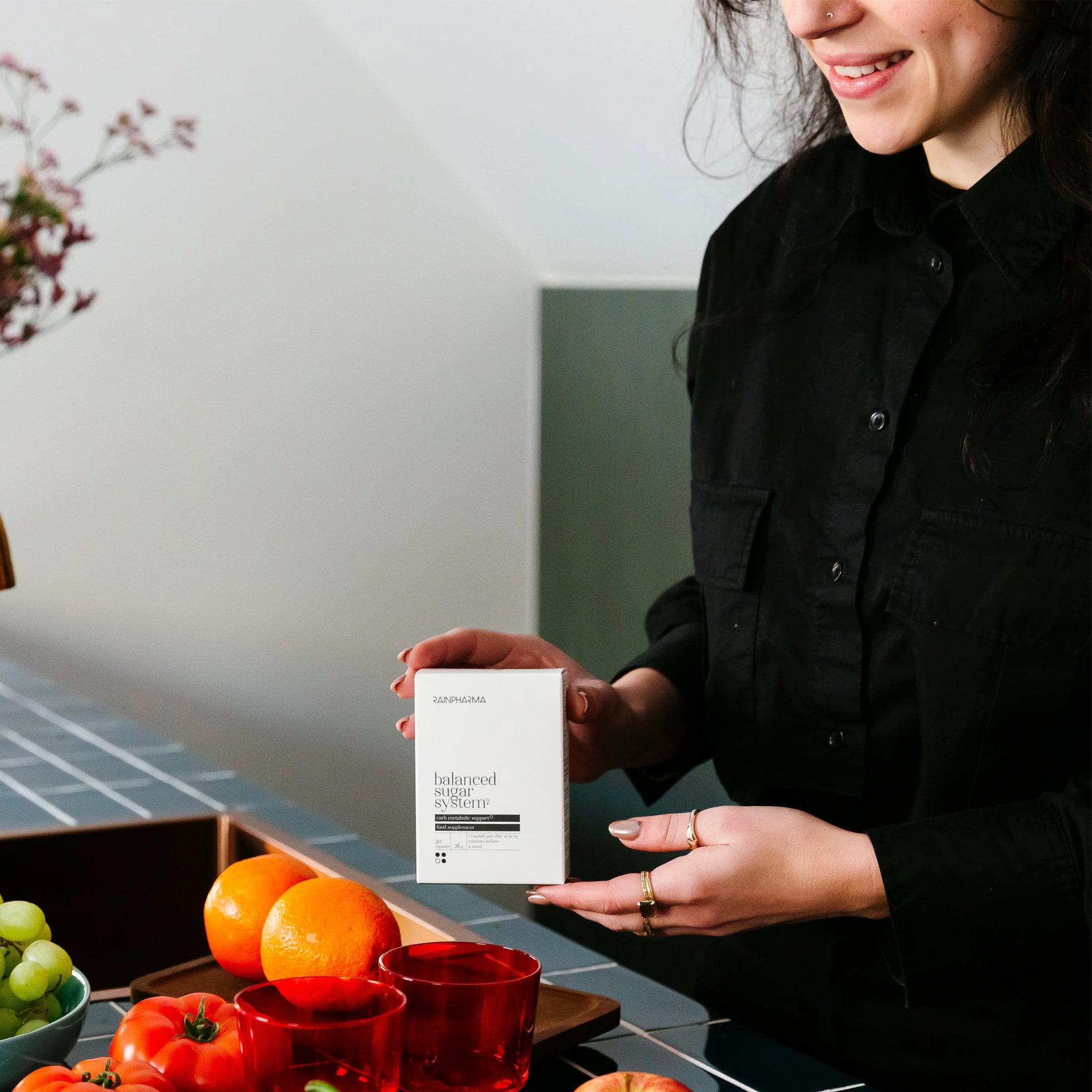 Een persoon in een zwart shirt houdt een doos vast met het label "Balanced Sugar System" van RainPharma. De tafel op de voorgrond is versierd met oranje, groene en rode glazen voor een impactvolle presentatie over bloedsuikerspiegel en darmgezondheid.