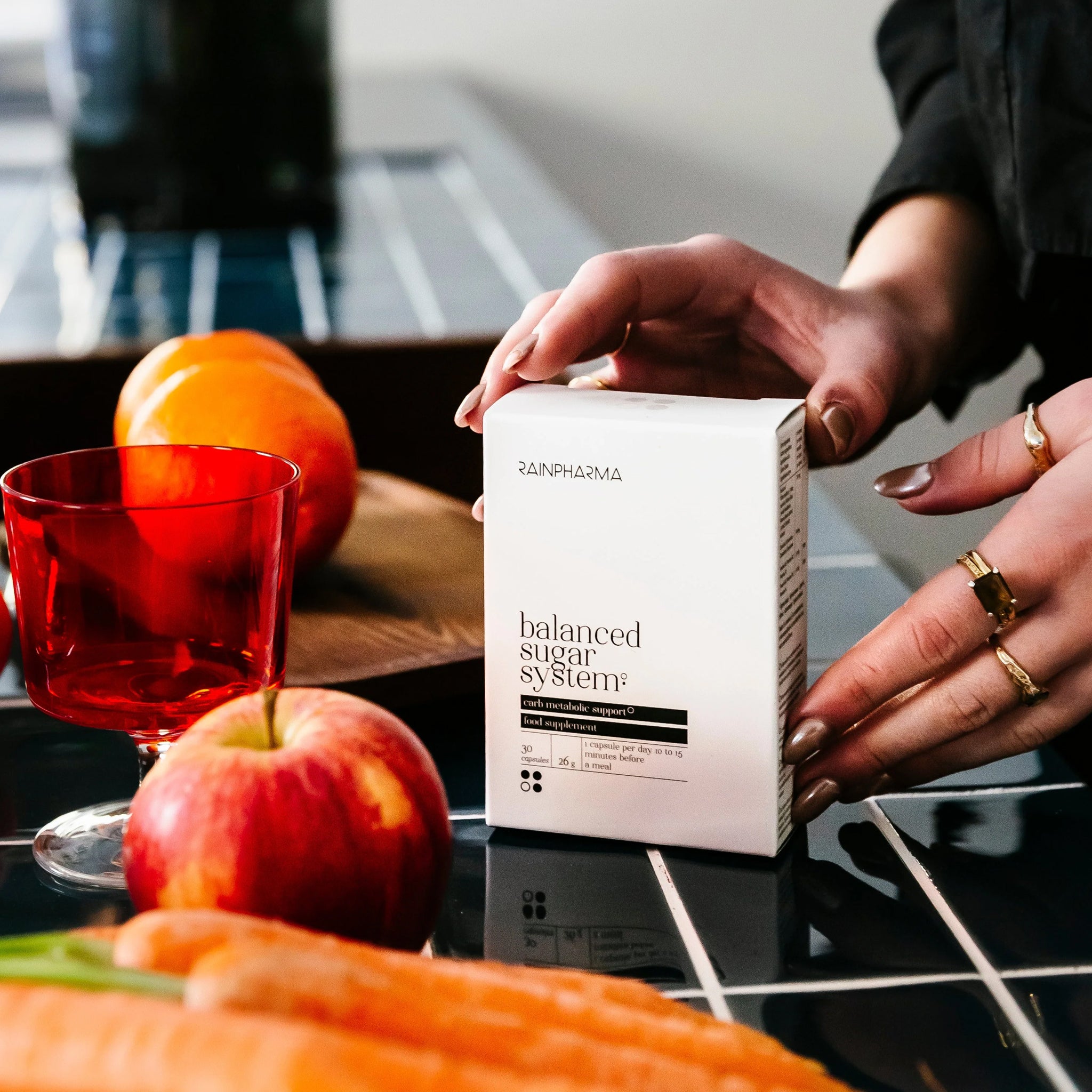 Een persoon houdt een doos vast met het label "Balanced Sugar System" van RainPharma, op een betegelde tafel met appels, wortels en een rood glas. Het product ondersteunt darmgezondheid en bevordert welzijn van binnenuit.