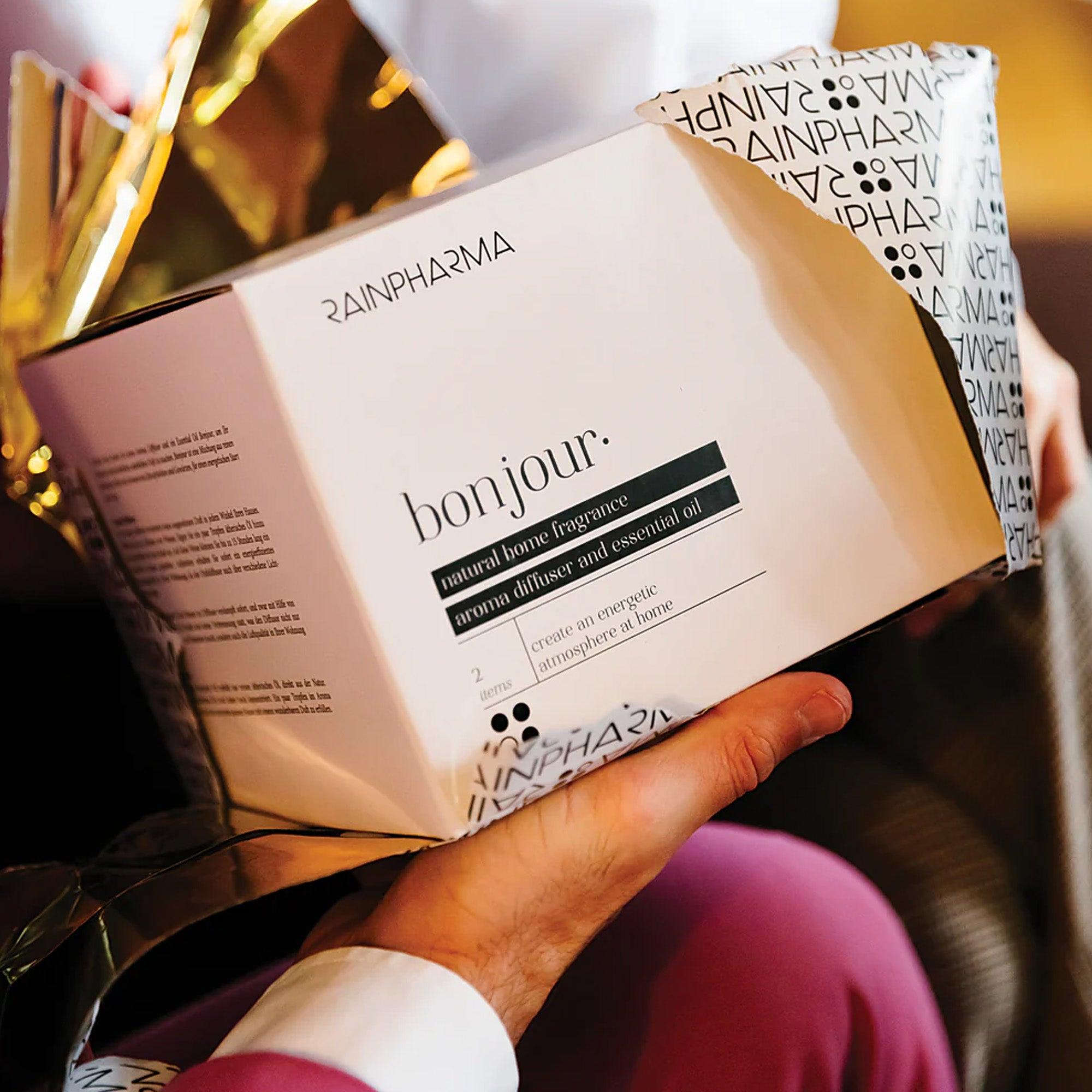 Een persoon houdt een ingepakte geschenkdoos vast met het opschrift "Aroma Diffuser Geschenkbox - Bonjour" van RainPharma. Hierin zitten een aroma diffuser en een flesje essentiële olie.
