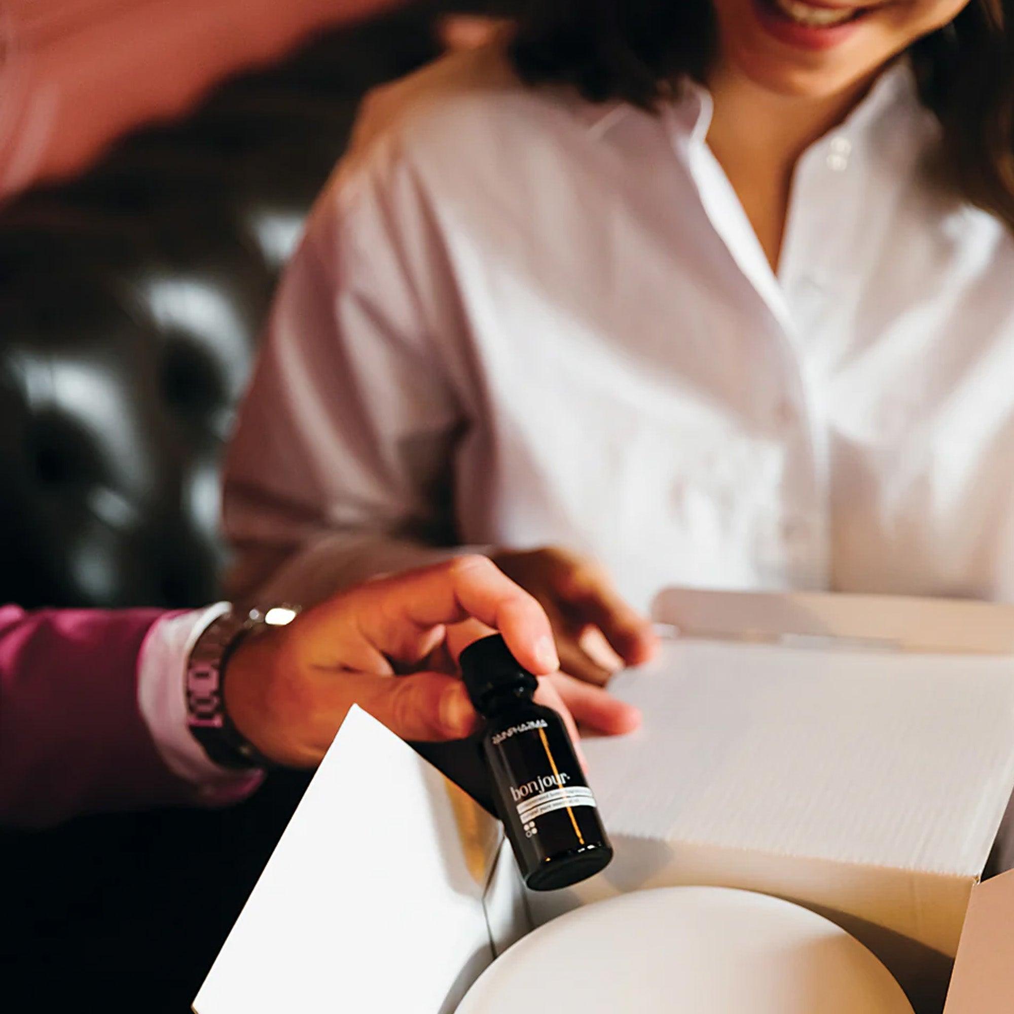 Iemand houdt een klein flesje essentiële olie vast bij een geopende Aroma Diffuser Geschenkbox - Bonjour van RainPharma, terwijl een ander persoon op de achtergrond glimlacht en waarschijnlijk geniet van de aangename effecten.
