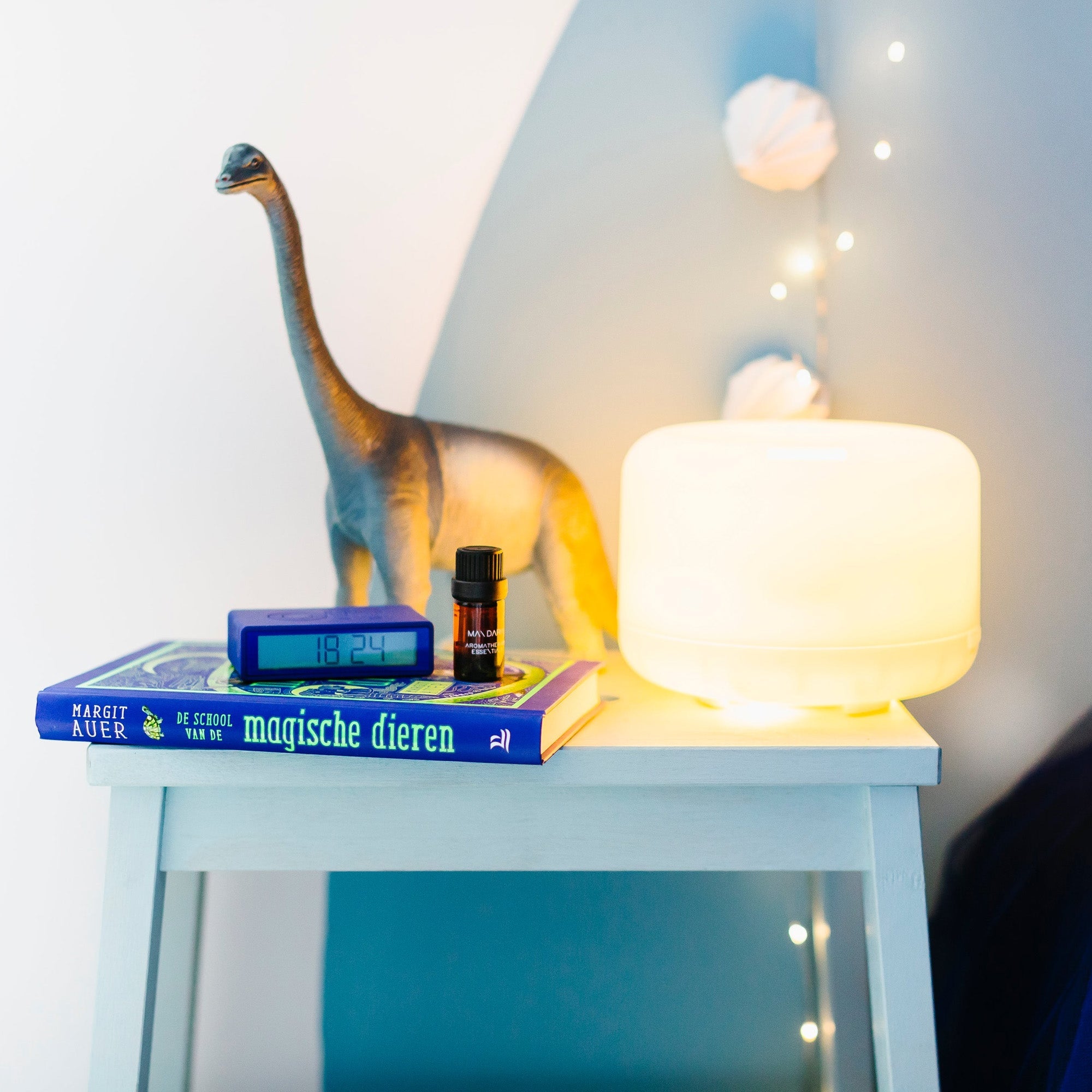 Een nachtkastje met daarop een klok die 18:24 aangeeft, een dinosaurusfiguur, twee boeken en een Aroma Diffuser Geschenkbox van RainPharma, samen met een gloeiende lamp, creëert een ontspannen sfeer die perfect is om te ontspannen.