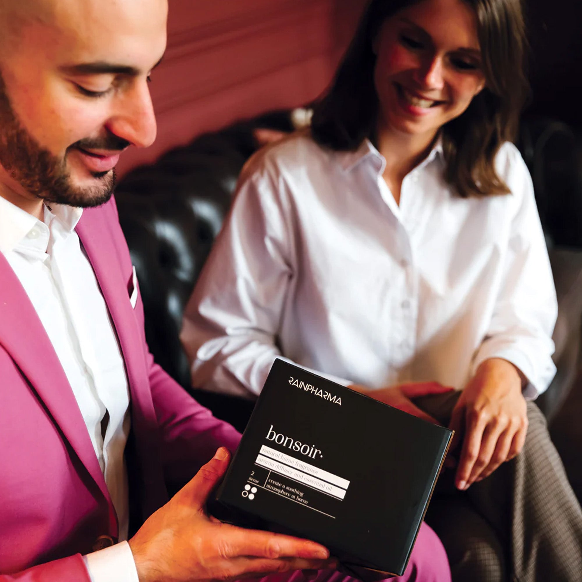 Op een donkere bank presenteert een man in een roze pak de Aroma Diffuser Geschenkbox - Bonsoir van RainPharma aan een lachende vrouw in een wit overhemd, terwijl een aroma diffuser de lucht vult met Ontspanning.