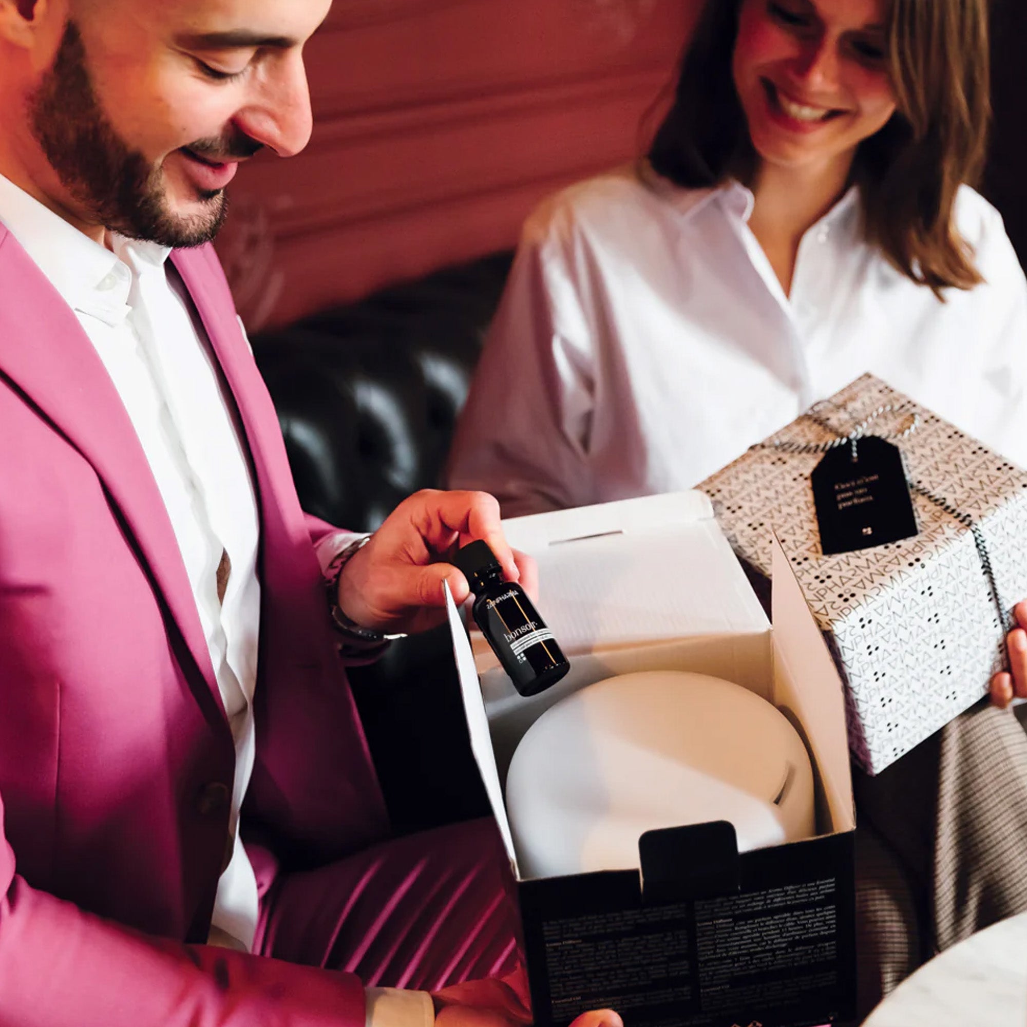 Een man in een roze pak presenteert de Aroma Diffuser Geschenkbox - Bonsoir van RainPharma, met een open geschenkdoos in zijn handen. Naast hem staat een glimlachende vrouw in een wit shirt, terwijl de rustgevende geur ontspanning en vreugde belooft.