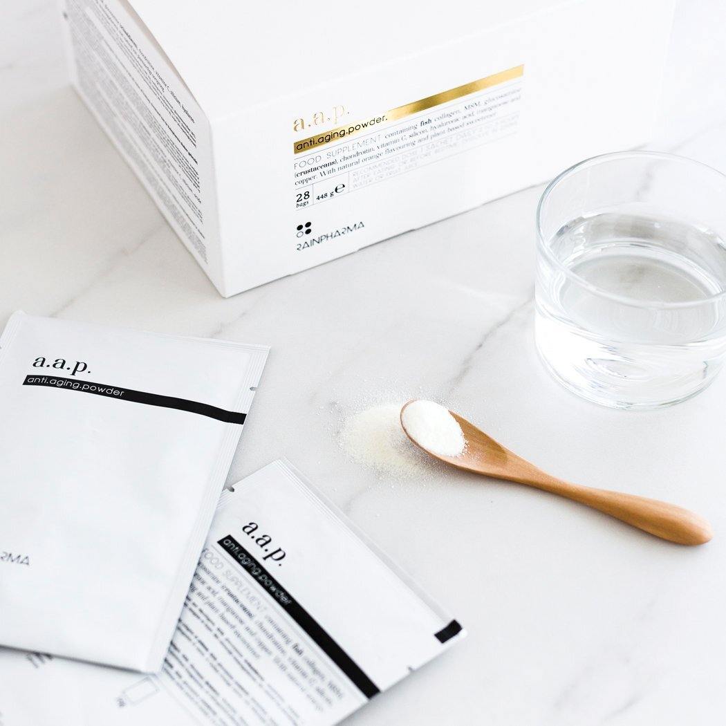 RainPharma Collageen Supplement - Pakjes anti-verouderingspoeder van AAP, een houten lepel met poeder, een glas water en de RainPharma productdoos staan op een wit oppervlak.