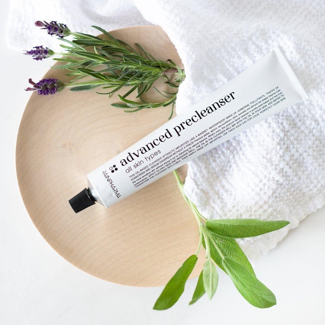 Een "Advanced Precleanser - gelaatsreiniger op oliebasis" van RainPharma voor de vette huid staat op een houten schaaltje, omringd door takjes groen en een witte handdoek, en bevordert zo een natuurlijke balans.