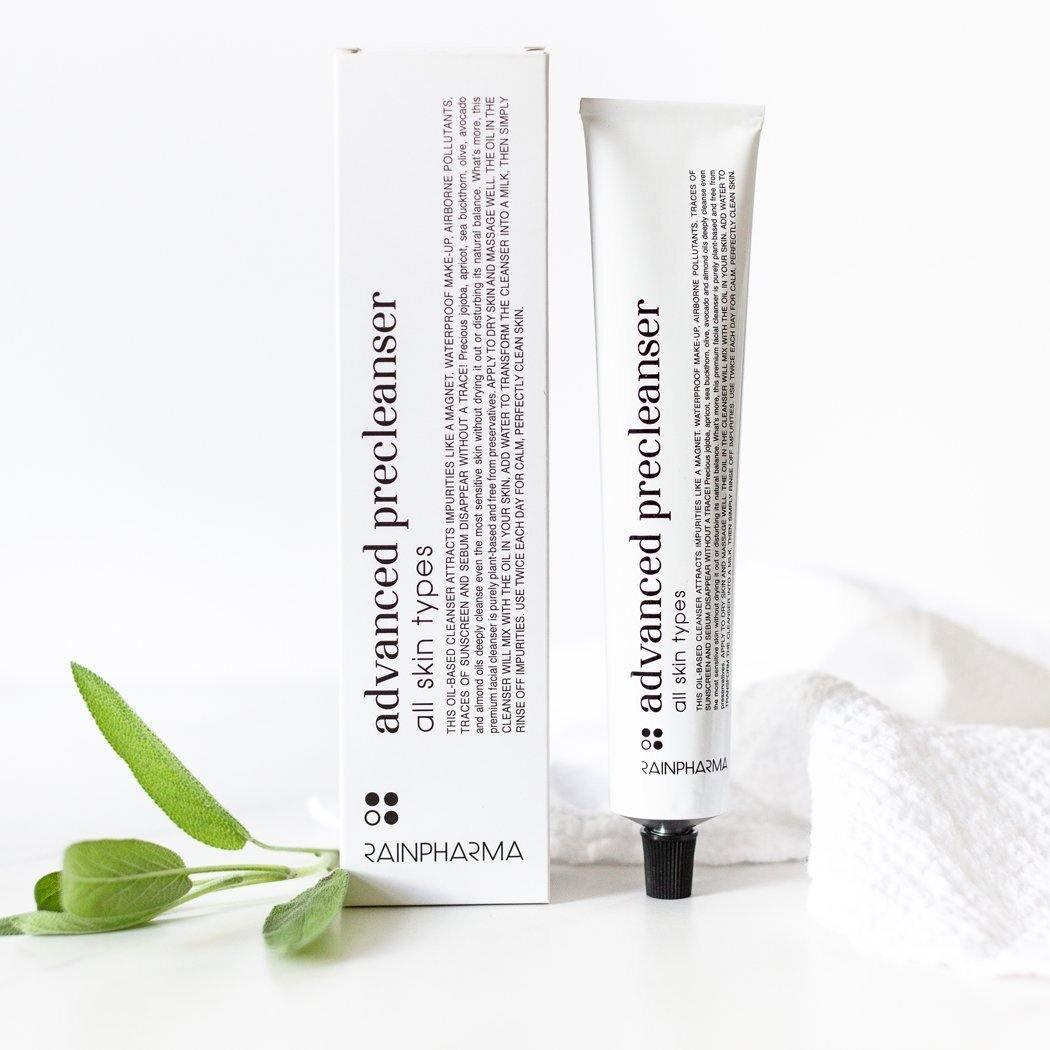 Naast de doos ligt een tube met het opschrift "Advanced Precleanser - gelaatsreiniger op oliebasis" van RainPharma, met een takje groene blaadjes en een witte doek voor een natuurlijke uitstraling.