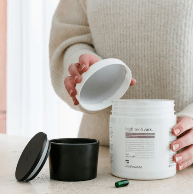 Een persoon opent een witte "High Tech AOX" container van RainPharma op een tafel, en onthult de rol ervan als voedingssupplement. In de buurt, een groene anti-oxidant capsule en zwarte deksel rusten, klaar om vrije radicalen effectief te bestrijden. Dit is de Family Pack met 365 capsules.
