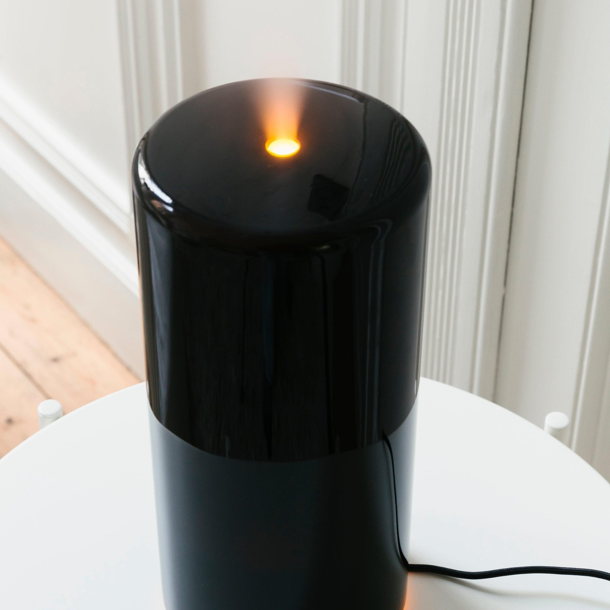 Een RainPharma Aroma Diffuser - Aurora Glass Sleeve in het zwart werpt een zacht oranje licht op de witte tafel en accentueert de lichtgekleurde muur met een sfeervol licht aan de bovenkant.